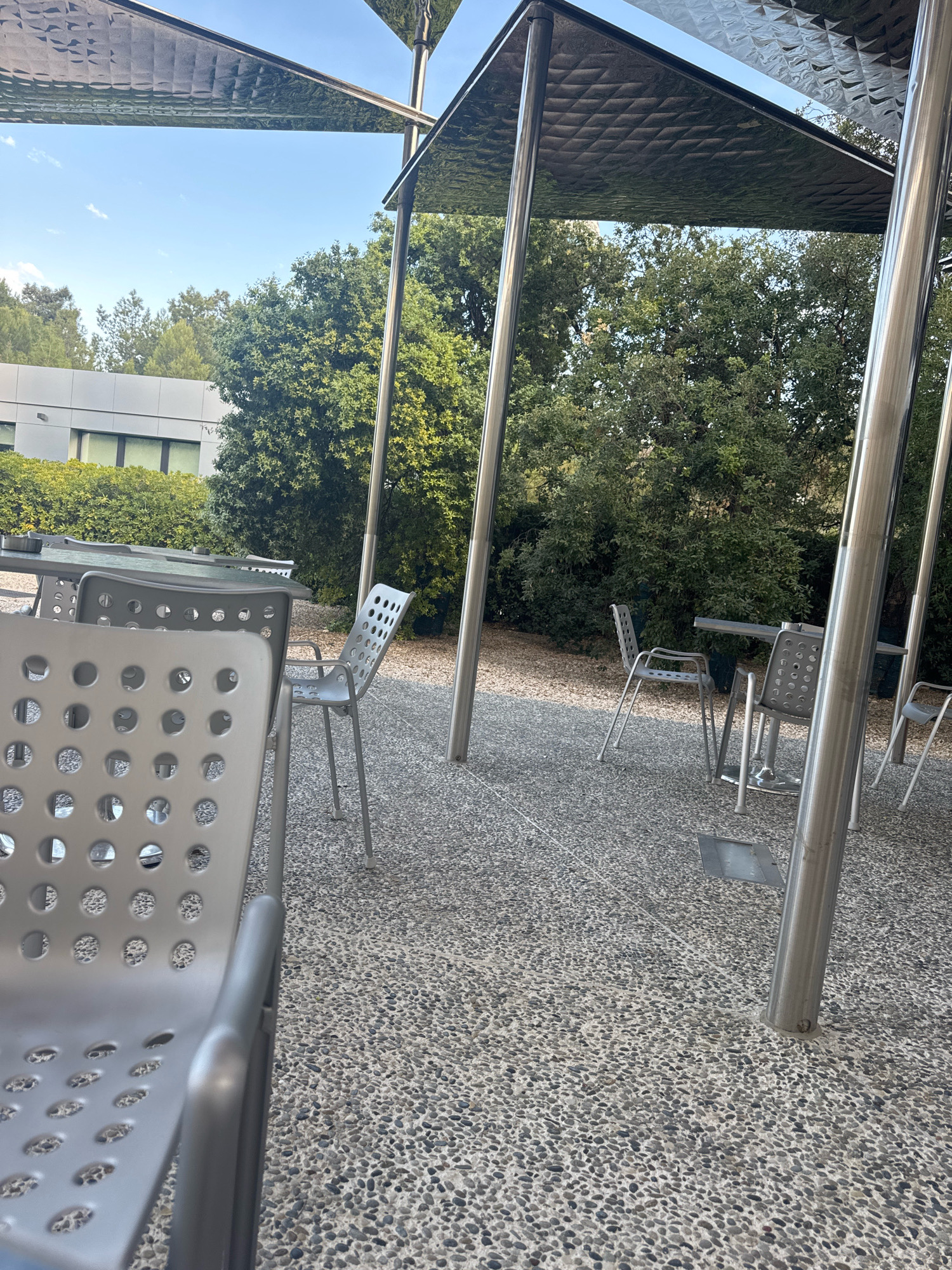 This image depicts an outdoor seating area under a modern, metallic canopy supported by vertical poles. The ground is paved with small, rounded pebbles. In the foreground, a grey, perforated chair is prominently displayed, its armrest curving into view. Several other grey metal chairs and tables are scattered throughout the scene, some partially obscured by the canopy's supports. Beyond the seating area, lush green trees form a dense backdrop, with a modern building visible in the distance to the left. The sky above is clear and blue. There are no people or animals present in the image. The scene appears to be a tranquil outdoor space, possibly a patio or a cafe seating area. The specific location in Strovolos, Cyprus is not discernible from the visual information provided.