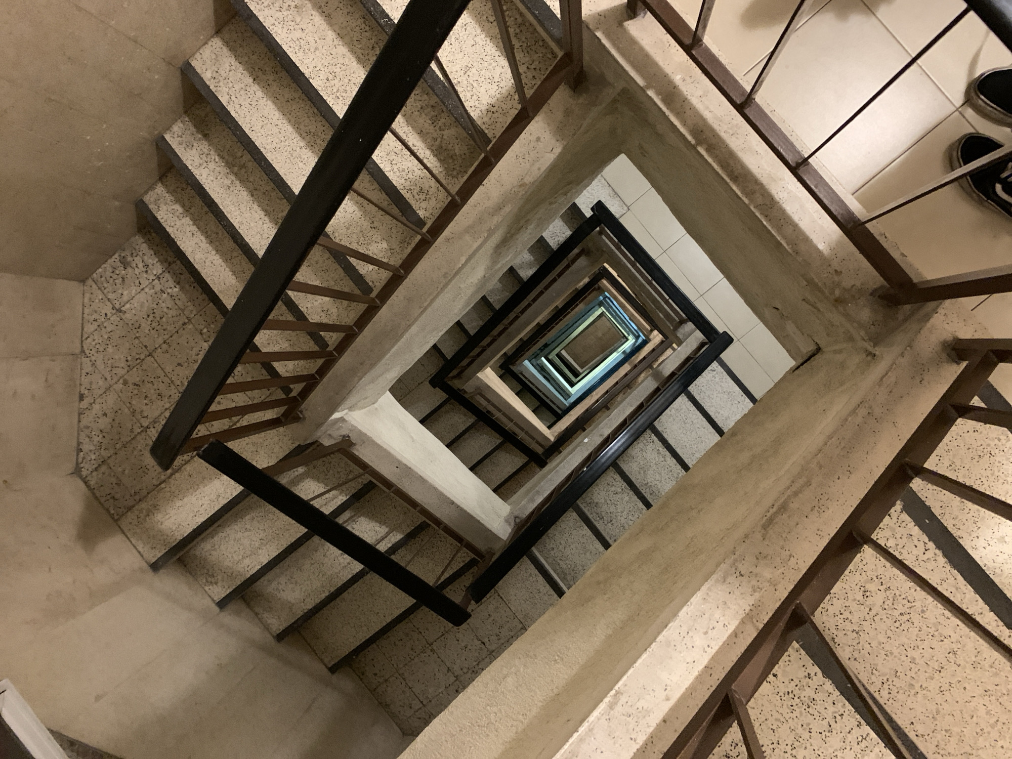 The image depicts a top-down view of a spiral staircase in what appears to be an interior space. The staircase is constructed of concrete steps with speckled light-colored tiling. Metal railings are present on either side of the staircase. Looking down the center of the spiral, a floor with light-colored tiles is visible. In the upper right corner of the frame, part of a shoe and leg are visible. There are no other visible people, actions, or interactions. The image lacks context clues to confirm a location of Tel Aviv, Israel.