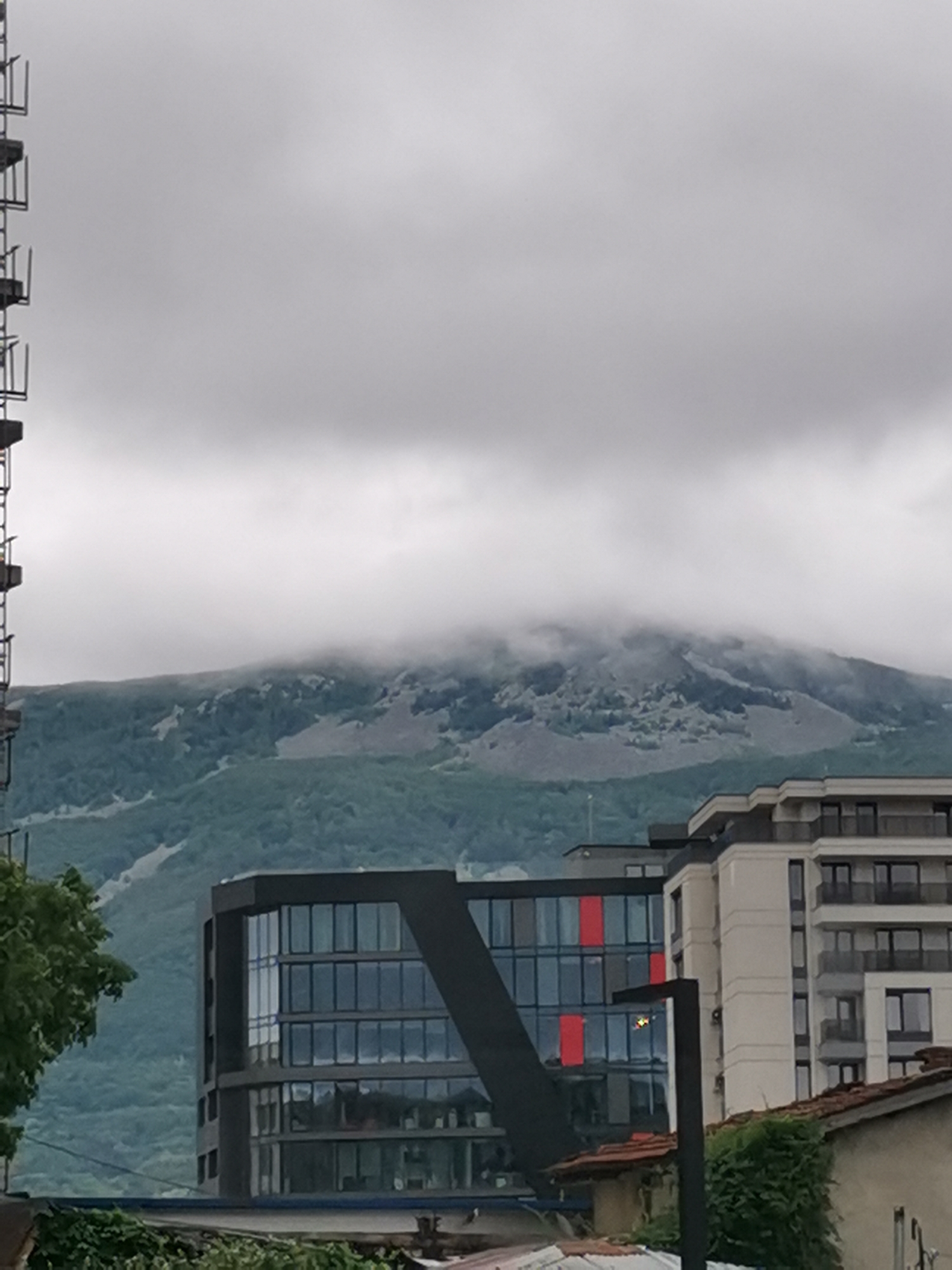 The image captures a view of a mountain range partially obscured by clouds, with modern buildings in the foreground. The mountain appears to be Vitosha Mountain, a prominent natural landmark situated on the outskirts of Sofia, Bulgaria. The sky is overcast, contributing to a diffused lighting condition.
In the middle ground, a modern office building with a distinctive angular design and a glass facade is visible. It has a dark-colored exterior with geometric accents and prominent windows reflecting the cloudy sky. Adjacent to it, a taller residential or office building with a lighter-colored facade and multiple floors with balconies is situated. Red panels are visible on the facade of the office building, adding a splash of color. A dark, modern lamppost stands in front of these buildings.
The lower foreground shows rooftops of older buildings, some covered with red tiles and greenery. A large tree with lush foliage is present on the left side of the image, partially obscuring the view of the office building. Construction scaffolding is visible on the far left edge of the image, suggesting ongoing development or maintenance in the area. The overall impression is a blend of urban development and the natural landscape of Sofia.
