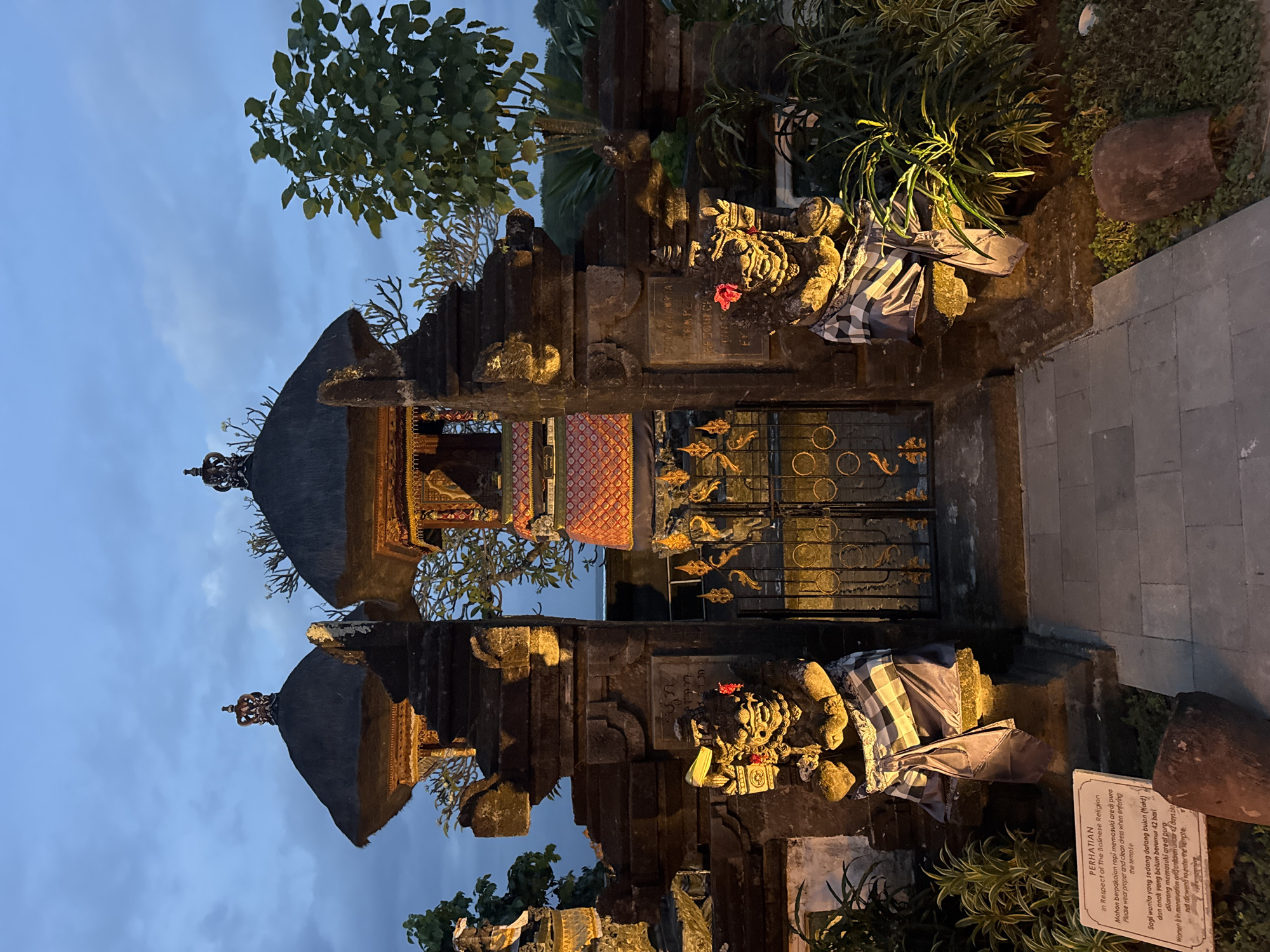 The image presents an exterior view of a traditional Balinese temple or religious structure. The structure is characterized by tiered roofs, decorative stone carvings, and ornate metalwork on the gate. Stone statues flank the entrance, each draped with a checkered textile, possibly a traditional sarong. Lush greenery surrounds the structure. The sky is dusky, suggesting it might be dawn or dusk. A sign is visible on the right. According to the OCR, the sign states "PERHATIAN In Respect of The Balinese Religion Mohon berpakaian rapi memasuki ared) pure Please wear proper and clean dress when entering the temple" and "Bagi wanita yang sedang datang bulan (Kald) dan anak yang belum berumur 42 hari dilarang memasuki areal pura Women is in menstruation grenildren uncur 2 days oto not allowend to enter the temple". Given the presence of the traditional structure and sign, the image suggests a location within a culturally significant area in Kuta Selatan, Indonesia, as indicated by the request.