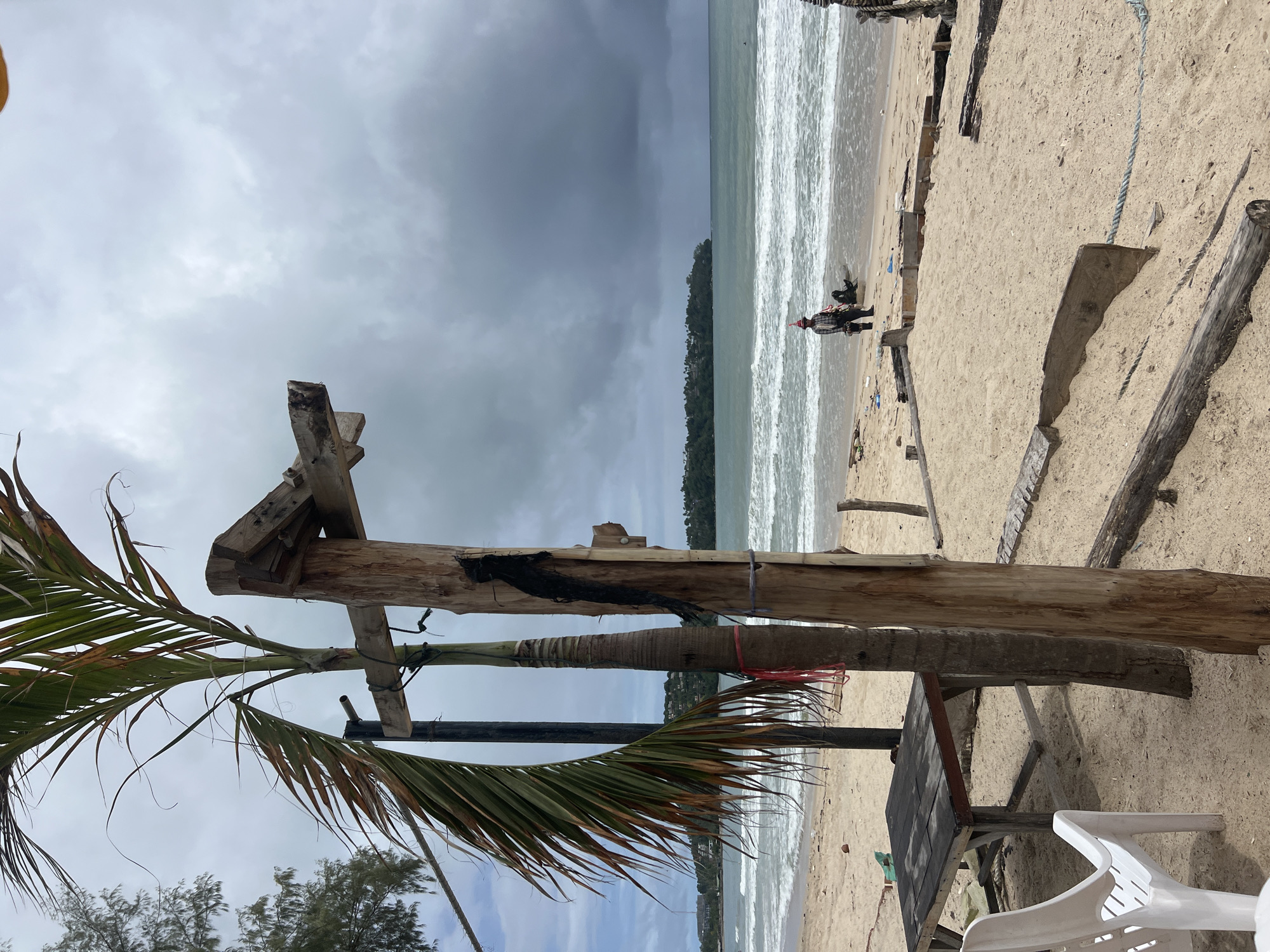 A vertical, medium shot captures a section of a sandy beach in Choeng Thale, Thailand, under an overcast sky. In the foreground, a rustic wooden structure, partially constructed and weathered, stands prominently. To the left, a mature palm tree with fronds showing some dryness leans into the frame, its trunk tied with red and blue twine. Nearby, a dark wooden table with visible wear and a white plastic chair are positioned on the sand. A short distance away, the ocean stretches to the horizon, its waves breaking gently on the shore. Two figures are visible on the beach near the water's edge. One individual, wearing a checkered shirt, dark pants, and a conical hat, stands facing away from the camera, while another figure is partially visible lying down near them. Scattered debris, including plastic bottles and driftwood, litters the sand. The background reveals a tree-lined coastline with buildings nestled among the foliage, suggesting the proximity of a town. The overall scene depicts a somewhat unkempt beach environment with signs of both natural elements and human activity.