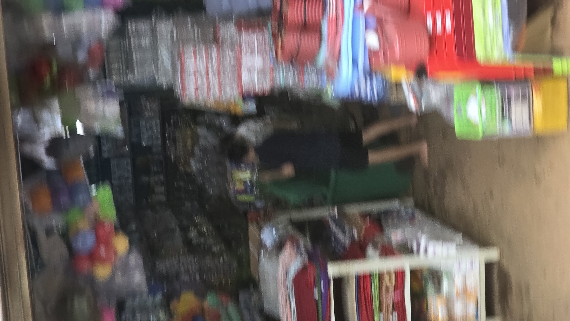 A cluttered store in Pơng Drang, Vietnam is filled with stacked plastic containers of various colors (red, blue, green, yellow) and assorted merchandise. A person wearing a dark shirt and shorts stands amidst the merchandise, near a green object. Shelving units display fabrics and other goods.