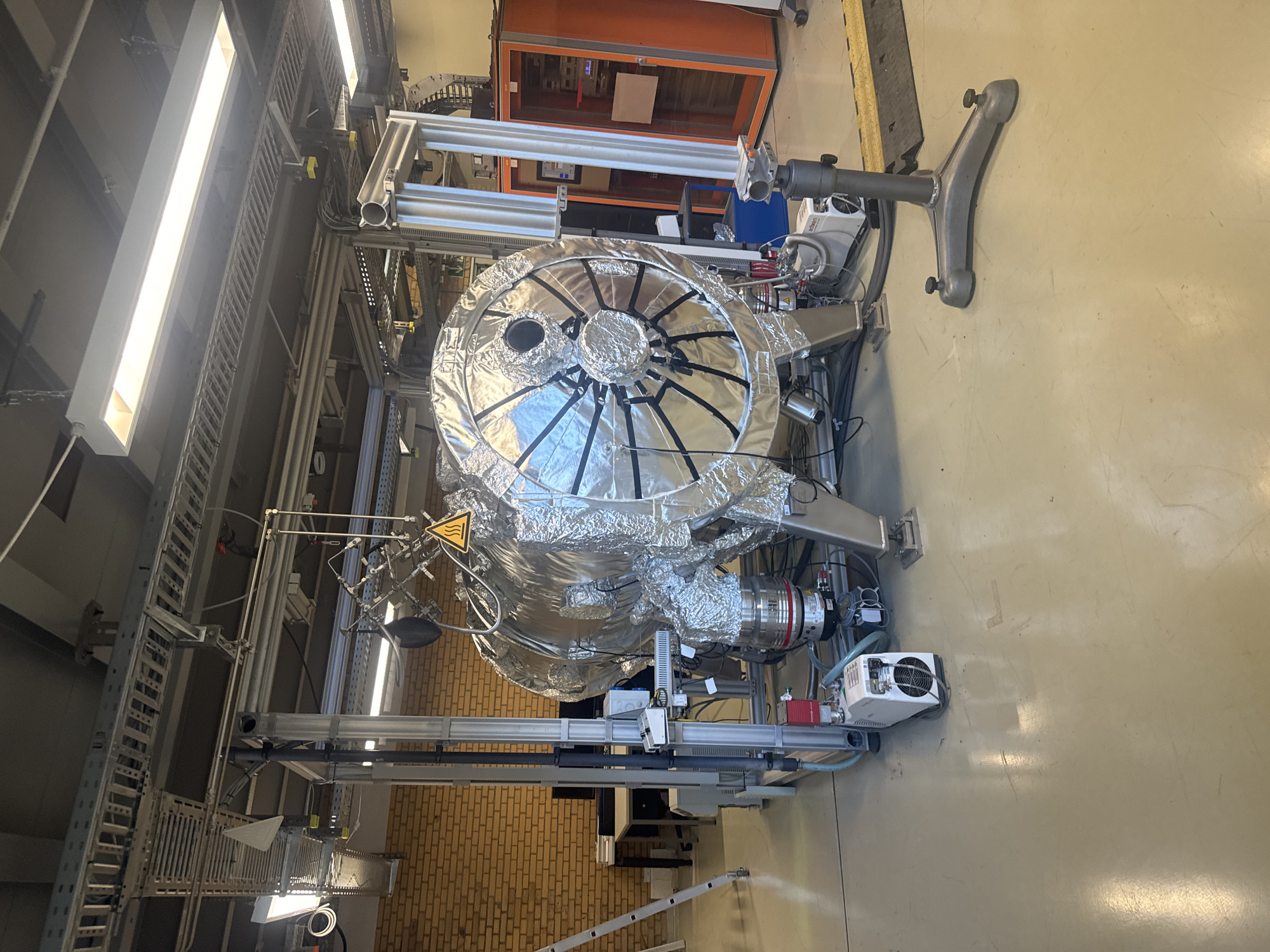 In a laboratory environment in Jülich, Germany, a large, cylindrical, silver-foil-wrapped vacuum chamber is the central focus. The chamber is supported by metal legs and connected to various pipes, wires, and smaller pieces of equipment, including a white unit with a fan and a red valve. A yellow triangular warning sign with a heat symbol is visible on the chamber. Overhead, a complex network of metal cable trays and lighting fixtures are mounted to the ceiling. To the right of the vacuum chamber, an orange electronic rack is present, containing various instruments and screens. A metallic tripod stands in the foreground to the right of the chamber. The floor is a polished, light-colored surface, reflecting the overhead lights. In the background, a brick wall is visible, along with a desk and computer monitor. A ladder is leaning against the wall on the left side of the image.