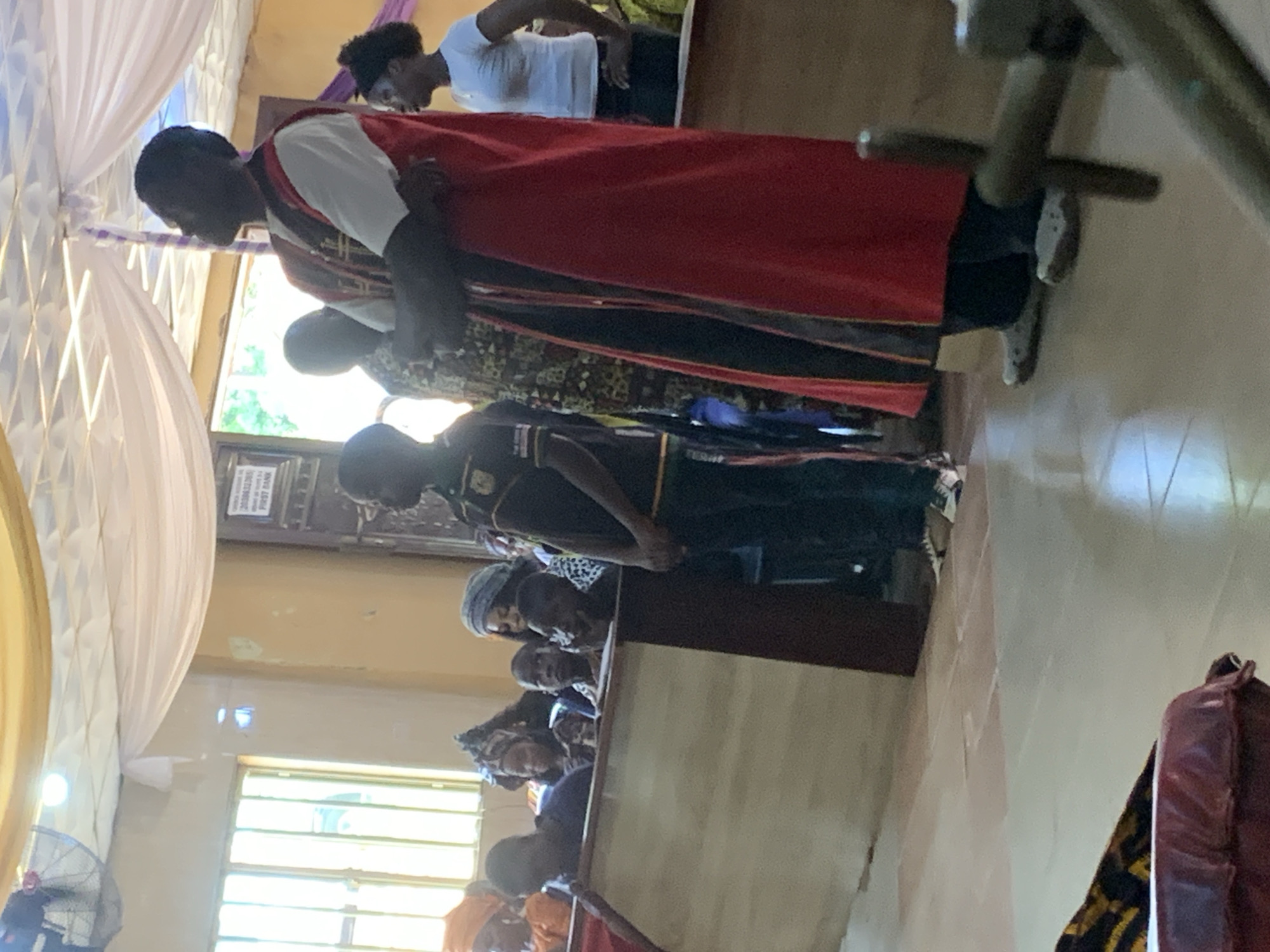 A group of people is seen indoors. A man in a white shirt and red robe stands with his head bowed, while others stand behind him. Additional people sit in what appear to be pews. The location seems to be inside a church or religious building, as evidenced by the architecture and the type of clothing worn by some individuals. The image does not confirm that this image was taken in Akpaka Forest Reserve, Nigeria.