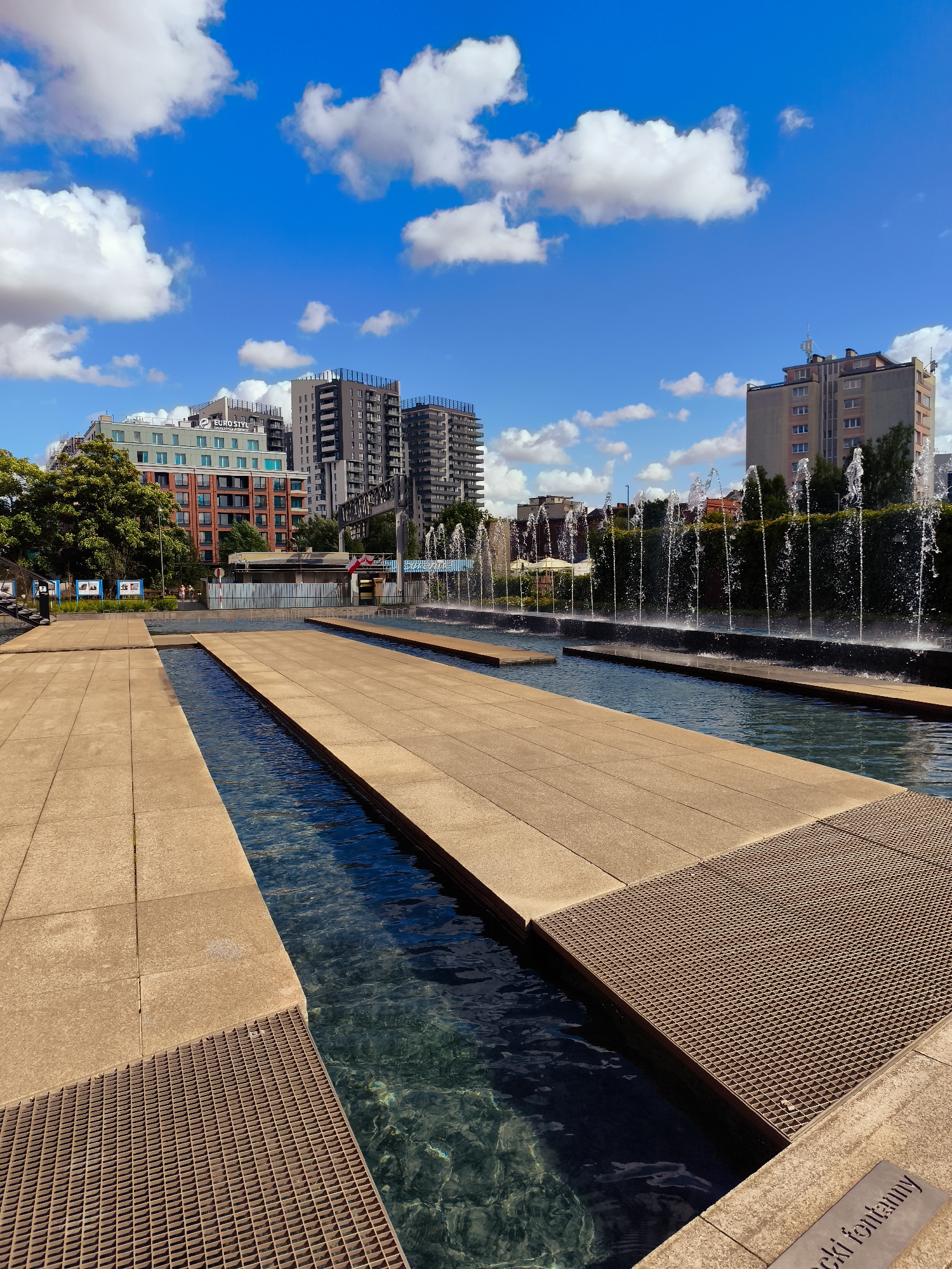 The image depicts the Pasażowe fountains in Gdańsk, Poland, on a sunny day with a bright blue sky and scattered white clouds. In the foreground, there are large, light-brown tiled platforms adjacent to shallow pools of clear, dark blue water. Metal grates are visible along the edges of the tiled areas. In the middle ground, a row of thin water jets sprays upwards from a dark, submerged line, creating a curtain of water against a backdrop of green bushes. Beyond the fountains, several modern buildings are visible, including apartment complexes and office buildings. One building on the left has the text "EURO STYL" on it. The overall scene is an urban public space designed with water features and contemporary architecture.