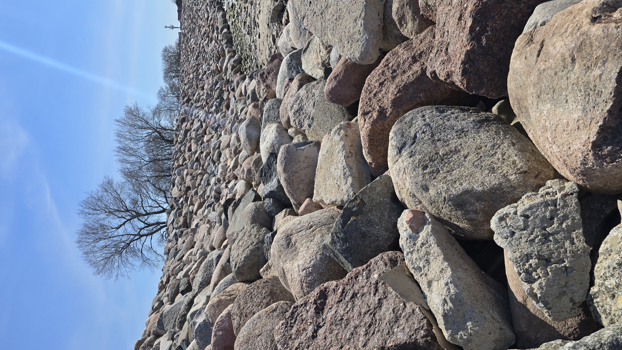 The image shows a wall constructed of various sized stones and boulders of mixed colors, predominantly tan, gray, and red. The wall extends diagonally from the lower right corner toward the upper left. Behind the wall and towards the upper left is a row of bare trees against a clear blue sky. A linear, bright light streak cuts through the sky. The location is identified as Baltiysk, Russia. There are no people visible in the image.