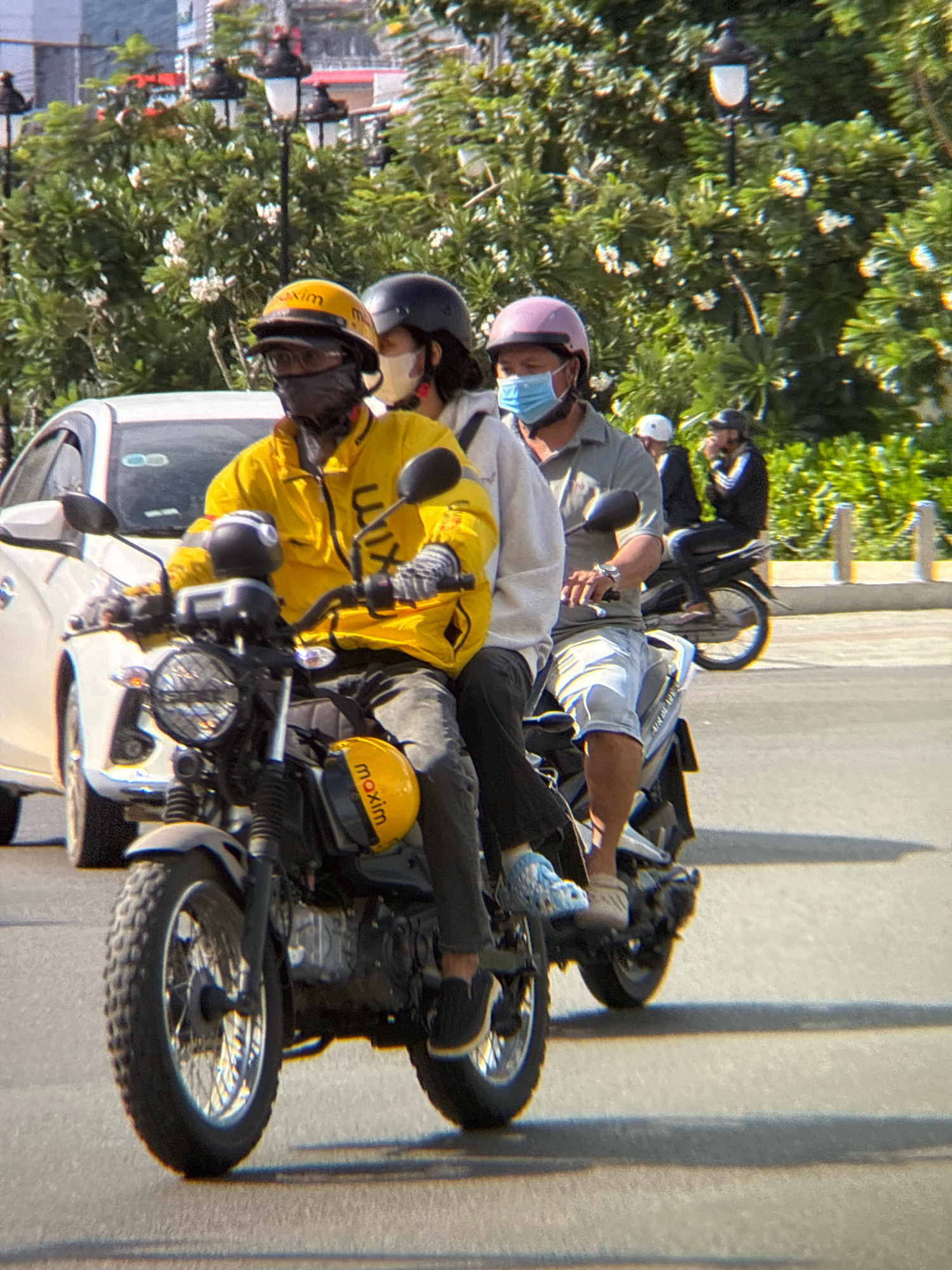 This image captures a busy street scene in Nha Trang, Vietnam, likely on a sunny day. In the foreground, three individuals are riding a single motorcycle, a common mode of transportation in the city. The rider in the front wears a yellow jacket with the "maxim" logo and a yellow helmet, along with a black face mask. Behind him, a passenger in a white jacket and a dark helmet is visible, also wearing a face mask. The third passenger, at the very back, is wearing a light gray shirt and a pink helmet, with a light blue face mask.
The motorcycle itself appears to be a scrambler-style bike with a black frame and chrome accents. The front wheel has a knobby tire, suggesting it's designed for varied terrain, though it's currently on an asphalt road. The background reveals lush green trees with white flowers, indicative of the tropical climate of Nha Trang. Streetlights are visible among the trees. To the left, a white car is partially visible, adding to the sense of urban activity. In the distance, other individuals on motorcycles and a building can be seen, further emphasizing the vibrant street life. The overall impression is one of everyday life in a bustling Vietnamese city.