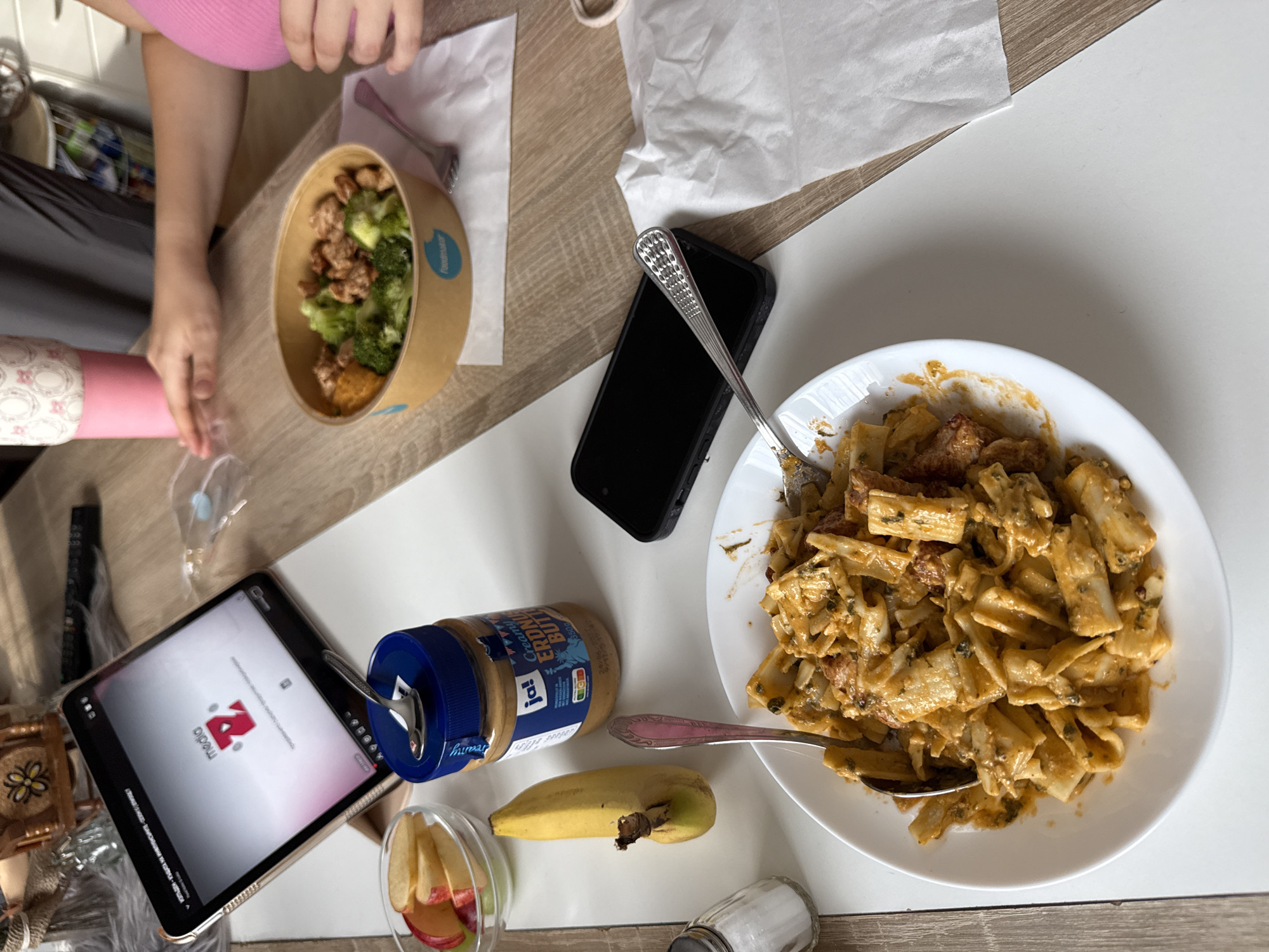 A close-up, high-angle, overhead shot captures a rustic wooden table laden with various food items and electronic devices. In the foreground, a white circular plate showcases a generous portion of pasta with a creamy orange sauce, interspersed with visible pieces of meat and herbs. Two silver forks rest on the plate, one partially submerged in the pasta. To the left, a small glass bowl contains sliced apples, alongside a whole banana and a salt shaker. A jar of creamy peanut butter with a blue lid is prominently placed, a spoon protruding from its opening. A dark smartphone lies on the table to the right of the peanut butter. Further back, a light brown takeout bowl holds a meal of chicken, broccoli, and a dollop of orange puree. A pink insulated cup stands to the left of this bowl, and a person's hands are visible in the background, one resting near the takeout bowl. A tablet computer, displaying a logo with the letters "media Z," is propped up on a stand in the upper left corner. The overall scene suggests a casual meal being enjoyed in a domestic setting. The specific location in Hanau, Germany is not ascertainable from the image.