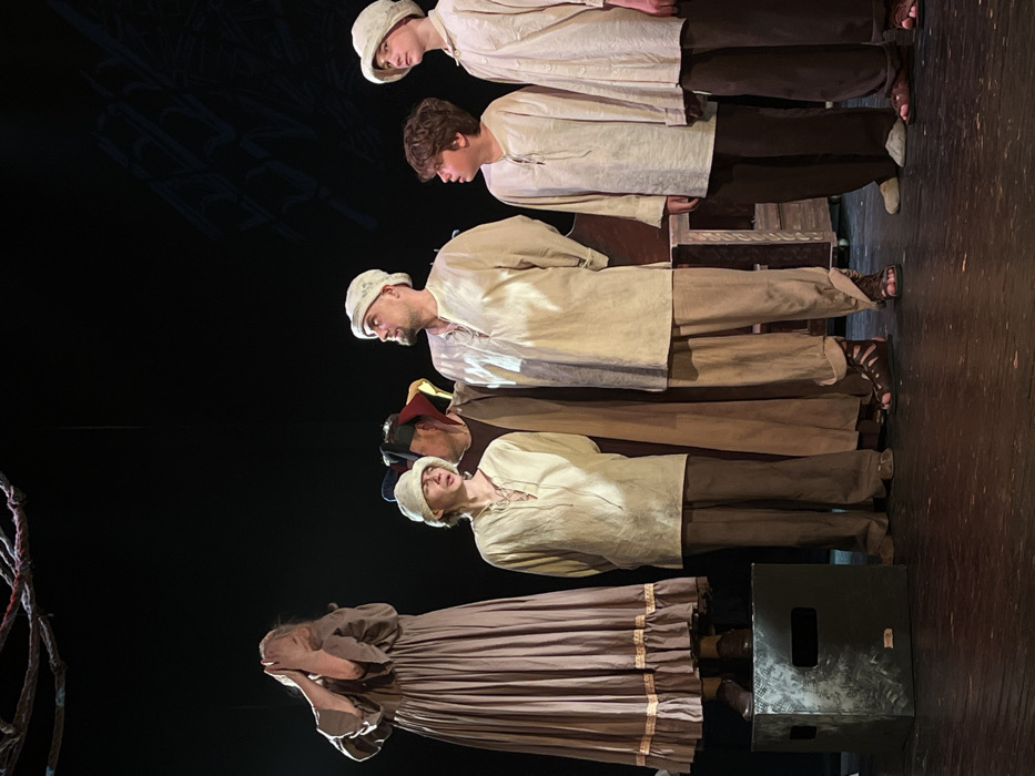 The image displays a theatrical performance on a stage, likely within a cultural center or theatre, located in Novoshakhtinsk, Russia. Six individuals, dressed in period-style costumes, are arranged across the stage. On the far left, a female figure in a long, flowing brown dress with ruffles stands atop a dark, cuboid stage prop, her hands covering her face in an gesture of distress. To her right, a male youth wearing a light beige tunic with laced neckline, dark trousers, and a light cap looks upwards and to the right. Adjacent to him, a partially obscured individual in a dark robe and a multicolored jester's hat is visible. At the center-right, a male adult, clad in a light beige tunic with laced neckline, matching trousers, and a light cap, stands with dark gladiatorial sandals, gazing upwards and to the right. To his immediate right, a male youth with dark, curly hair, dressed in a light beige tunic and dark trousers with light slippers, looks left towards the central adult. On the far right, a final male youth, wearing a light beige tunic, dark trousers, and a light cap, also looks upwards and to the right, wearing dark sandals. The stage features a dark background with abstract, branch-like suspended elements overhead and faint geometric patterns on the back wall, along with a partial wooden furniture prop on the right.