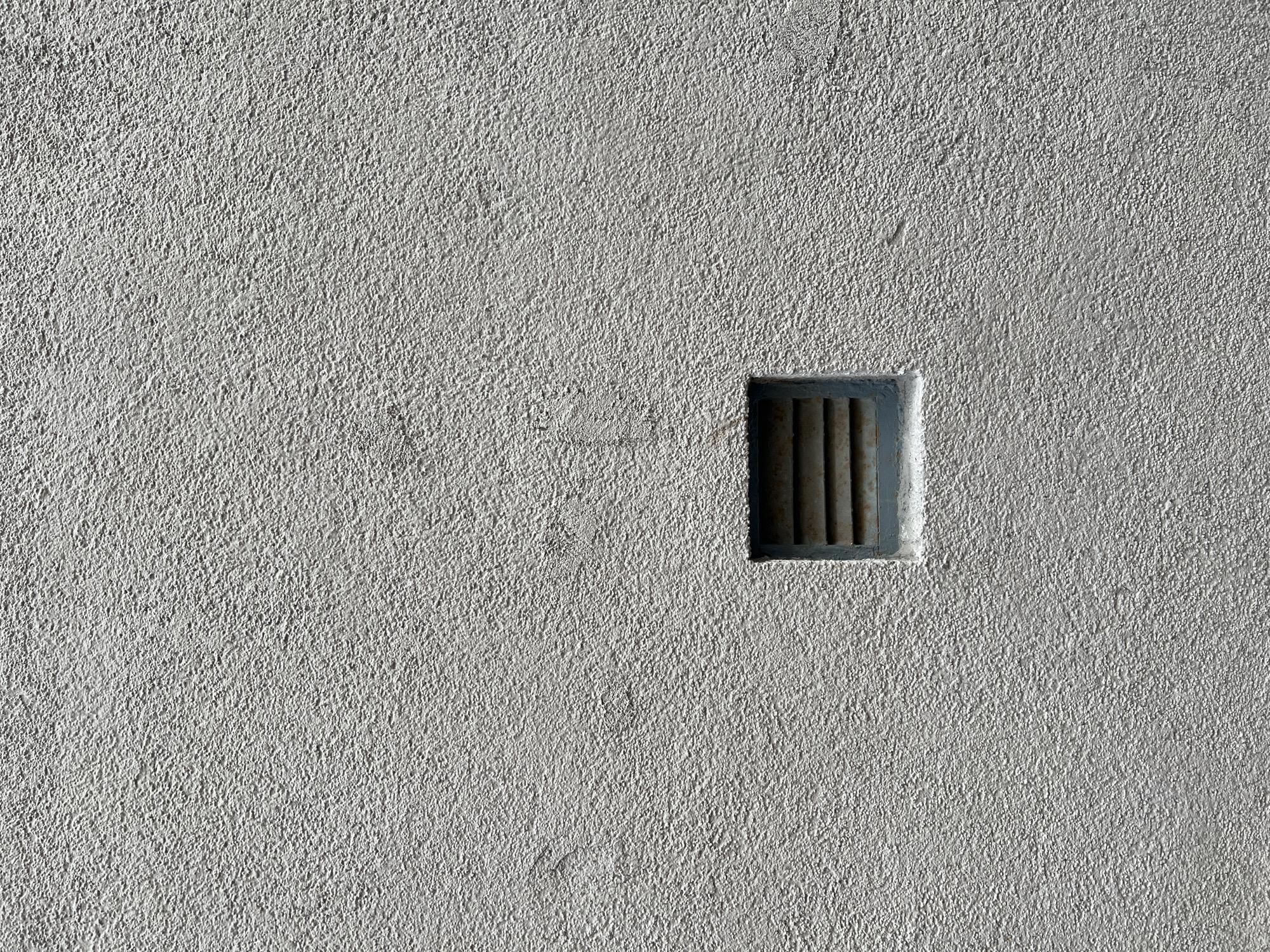 A close-up shot features a section of a light gray, heavily textured wall. Embedded within the wall is a small, square opening protected by vertical bars. The location, Saint Petersburg, Russia, is not discernible from the visual elements provided. No people, specific subjects beyond the wall and vent, actions, or interactions are visible. The focus is strictly on the texture and structure of the wall and the embedded vent.