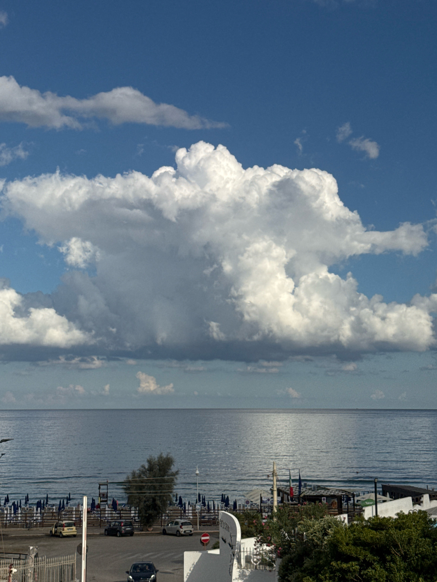 The image depicts a daytime view of the sea and a beach in Capaci, Italy. The sky is blue with a large, prominent cumulus cloud formation dominating the upper half of the frame. Below the sky, the calm sea stretches to the horizon. Along the shoreline, a beach is set up with rows of blue umbrellas and lounge chairs. A small tree stands near the center of the frame, with a few cars parked in a lot in front of the beach access. Buildings and foliage are visible on the right side of the image. The overall scene suggests a peaceful coastal setting under a partly cloudy sky.
