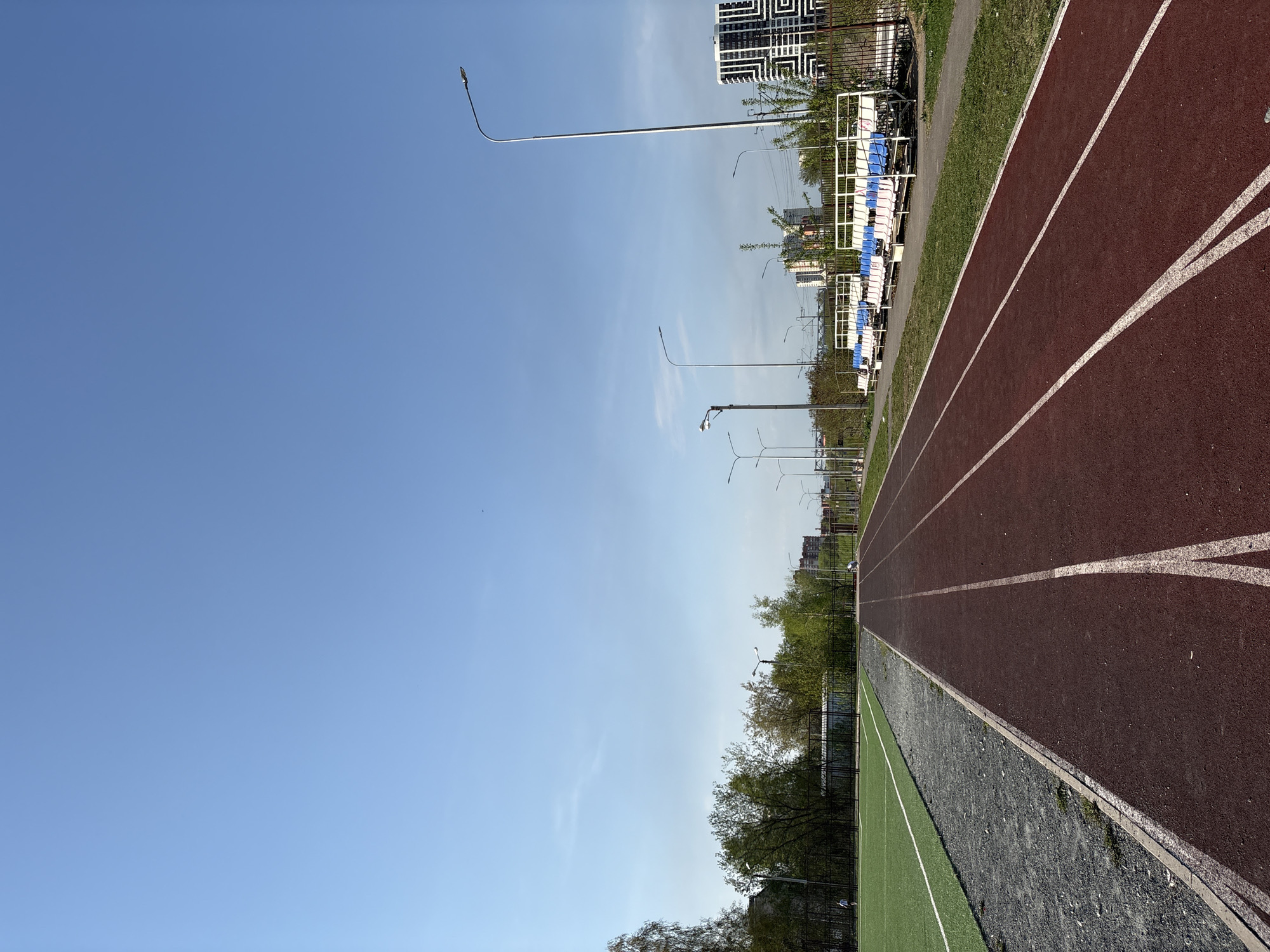 The image depicts an outdoor athletic track, likely in Perm, Russia, based on context. The track has a red running surface with white lane markings. An area of green turf and black gravel borders the track. Streetlights line the area beyond the track, and bleachers with white and blue seating are visible. A modern building is also in the background. The sky is blue with sparse clouds.