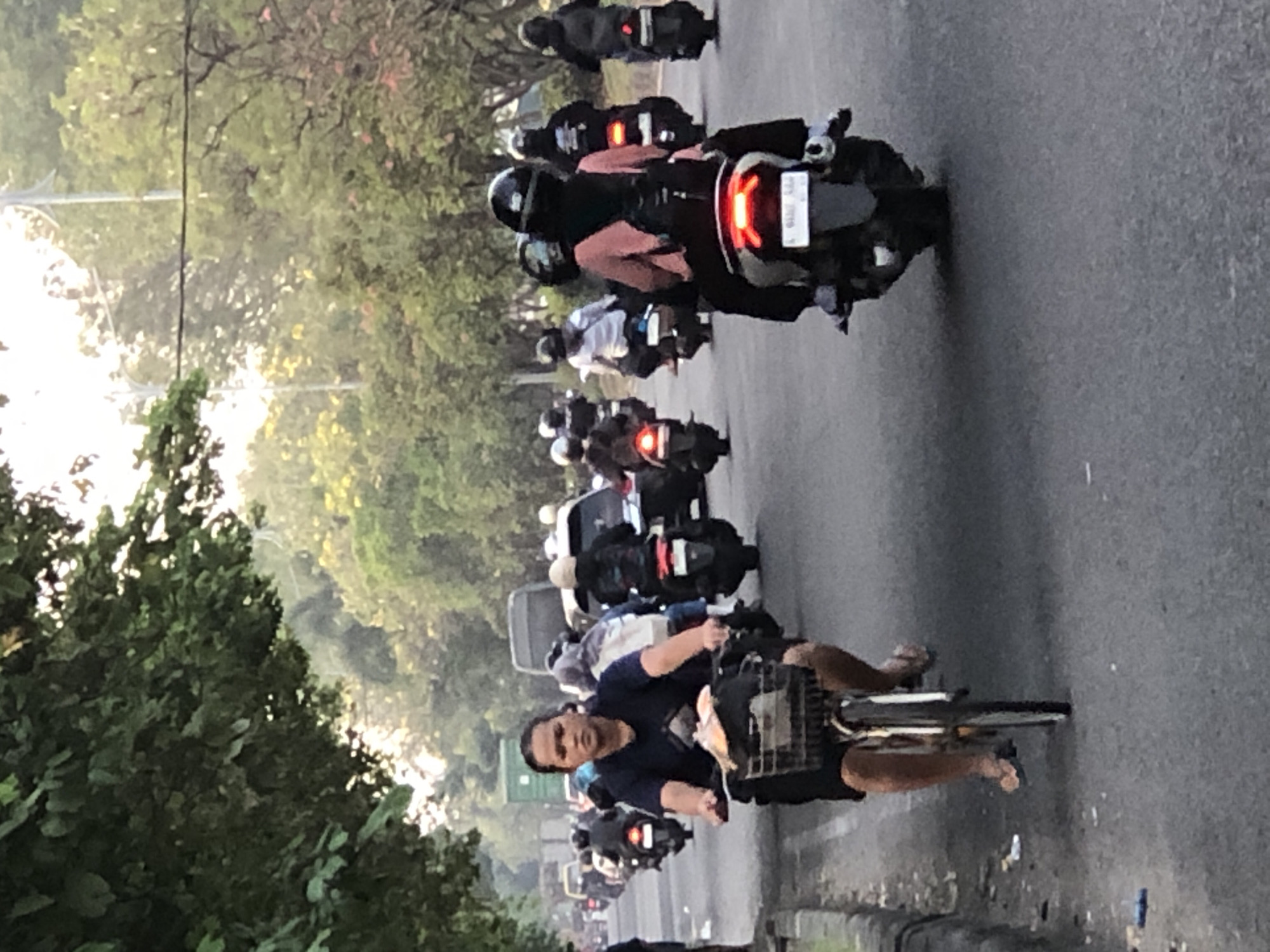 The image depicts a street scene in Sumorame, Indonesia, likely during the day with overcast skies or early morning/late afternoon light. A woman is riding a bicycle in the foreground, wearing a dark blue t-shirt and black shorts, with a basket attached to her handlebars containing some items. She is looking forward, and her feet are in flip-flops. Behind her, a convoy of motorcycles is visible on the road. Many of the motorcyclists are wearing helmets, with some passengers also present. The vehicles appear to be traveling in the same direction as the cyclist. Trees line the right side of the road, and there is a curb on the left side. The road surface is asphalt, with some debris visible. In the background, there are more vehicles and foliage, suggesting a moderately busy urban or suburban environment. A large green shipping container is visible on the left side of the road in the distance.