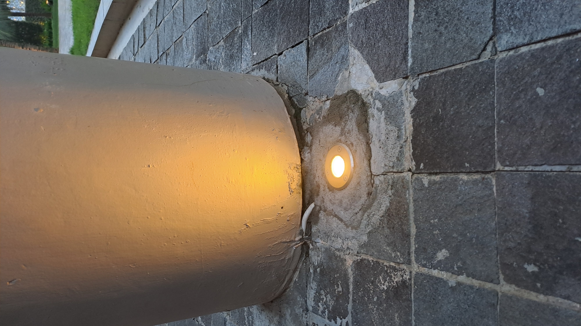 A low-angle, close-up outdoor shot focuses on the base of a large, light-colored cylindrical planter situated on a dark gray stone-tiled pathway. The planter is illuminated from its lower left by a warm yellow light source, casting a soft glow on its textured surface. Embedded in the pathway directly in front of the planter is a circular, flush-mounted light fixture emitting a bright white light. A white cable exits from the base of the planter and lies on the ground, leading towards the pathway. The stone tiles are weathered and uneven, with some cracks and areas of discoloration. In the background, to the right, a strip of green grass and a hint of palm trees are visible, suggesting an outdoor setting in a tropical or sub-tropical environment, potentially a resort or landscaped area in Daroykameu, Indonesia. No people or animals are present in the frame.