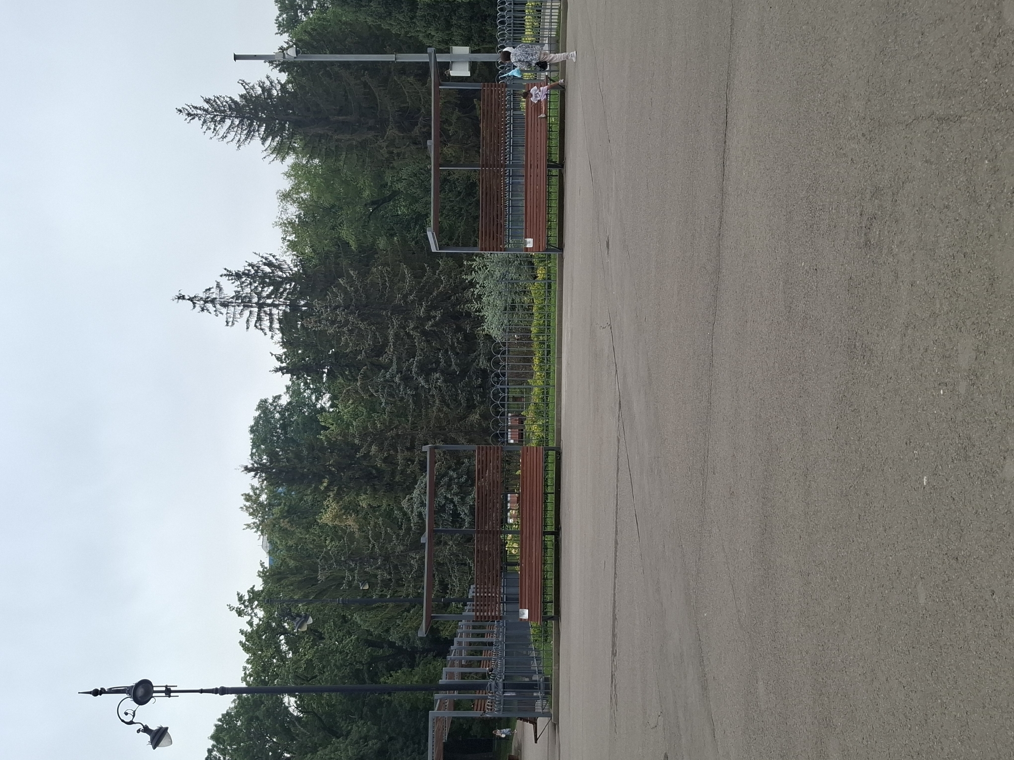 The image shows an overcast day in a park in Almaty, Kazakhstan. In the foreground, there is a wide expanse of asphalt pavement. In the middle ground, there are two identical covered benches with wooden slats. The bench on the right has a woman standing near it, and a young child sitting on the bench. Behind the benches is a decorative metal fence with a floral pattern, and beyond that, a dense line of trees, including tall coniferous trees and deciduous trees. A street lamp with two lights is visible on the left side of the image, and a security camera is mounted on a pole on the right side, partially obscured by trees. The sky is a uniform light gray.