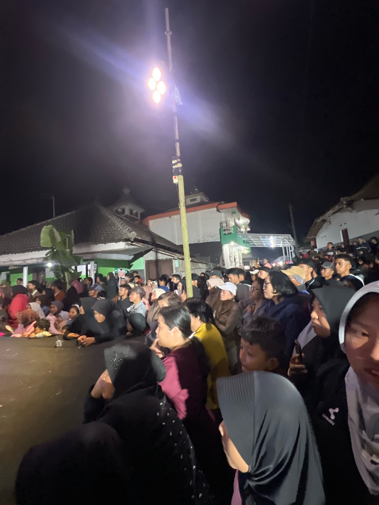 This image captures an outdoor night scene in Madyocondro, Indonesia, likely during a community gathering or event. A prominent floodlight on a pole illuminates a large crowd of people of various ages. The crowd is densely packed, with many individuals sitting or standing. In the background, traditional Indonesian houses with dark tiled roofs are visible, along with a building that appears to be a mosque or a similar religious structure with a green roof and white walls. The overall atmosphere suggests a lively and communal occasion, possibly a religious ceremony, festival, or social event taking place in the village. The lighting creates a stark contrast between the illuminated area and the dark night sky.