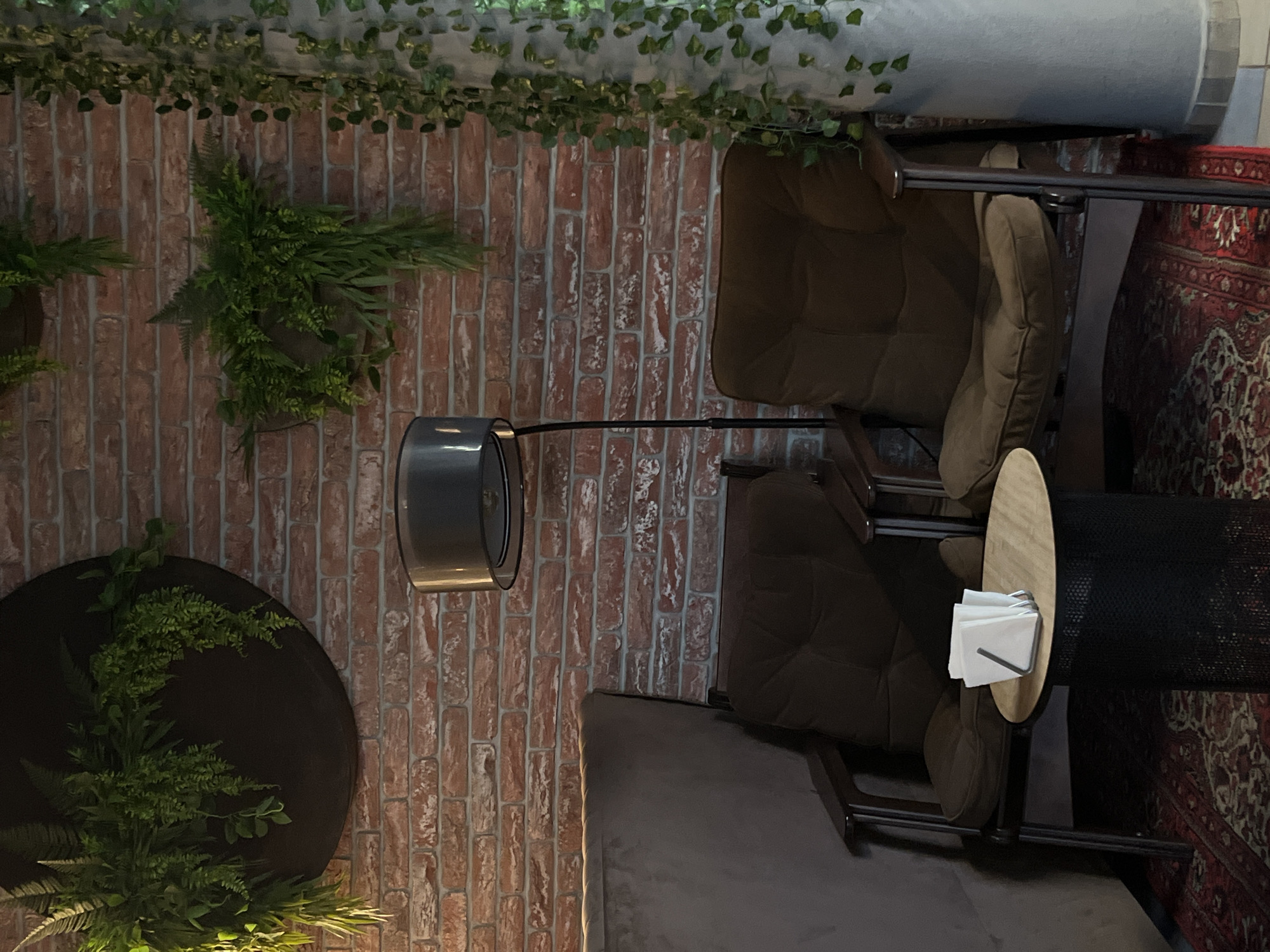 The image shows a lounge area with two brown armchairs, a small round side table with napkins, and a dark mesh-like container next to the table. A floor lamp with a grey shade stands between the chairs. The backdrop is a brick wall adorned with hanging plants and greenery. A patterned rug is partially visible in the corner. The location is likely a cafe or waiting area, potentially in Neftekamsk, Russia, though no specific details in the image confirm this. There are no people visible.