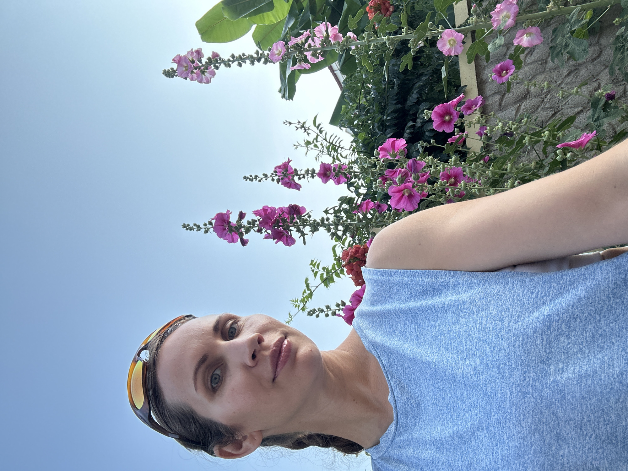A woman wearing a blue tank top and sunglasses pushed up on her head stands in front of a wall with pink hollyhocks and green foliage. She is looking towards the camera. The background shows a clear blue sky and the upper portion of a building. The scene appears to be outdoors, possibly in a garden or yard.