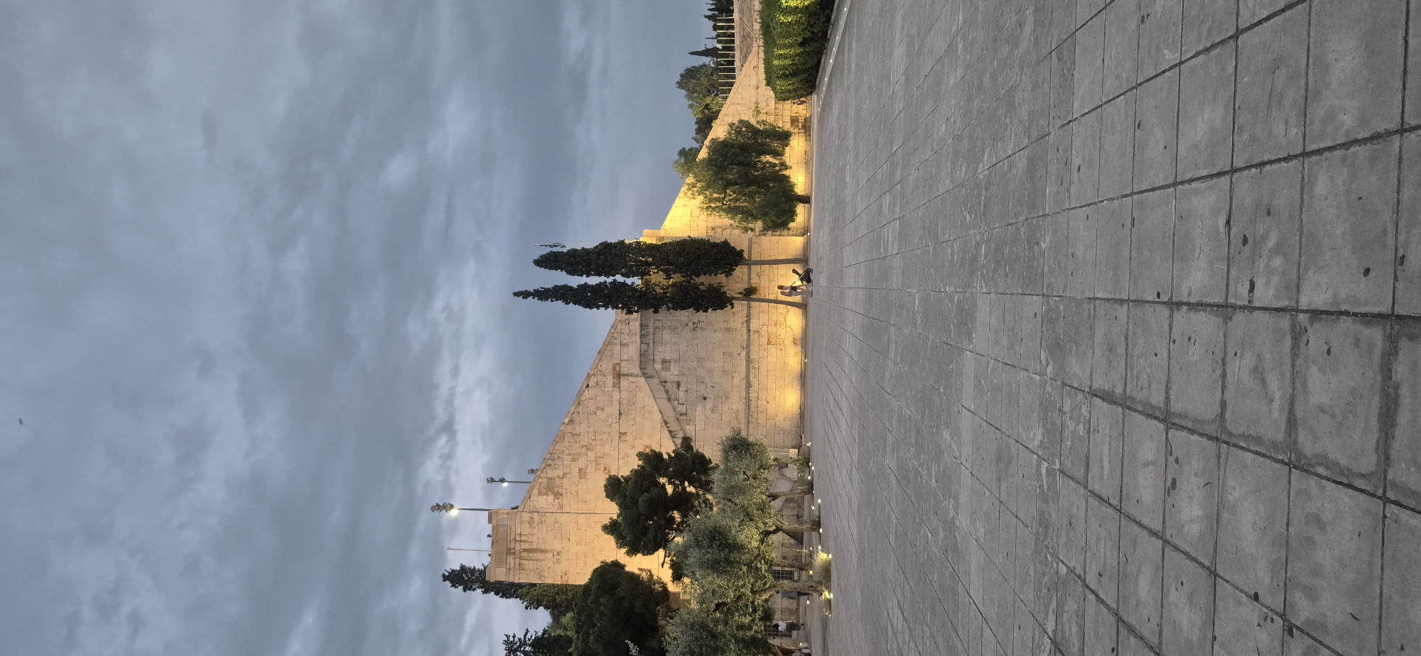 The image shows the Panathenaic Stadium in Athens, Greece, at dusk. The stadium's marble facade is illuminated by warm lighting, creating a stark contrast with the overcast sky. The foreground is a vast, tiled plaza. In the mid-ground, several trees, including olive trees, flank the stadium, with some smaller lights embedded in the ground around their bases. Two people are visible sitting on the stadium steps, and another person is walking with a stroller. The overall atmosphere is calm and serene, with the dimming light suggesting the end of the day.