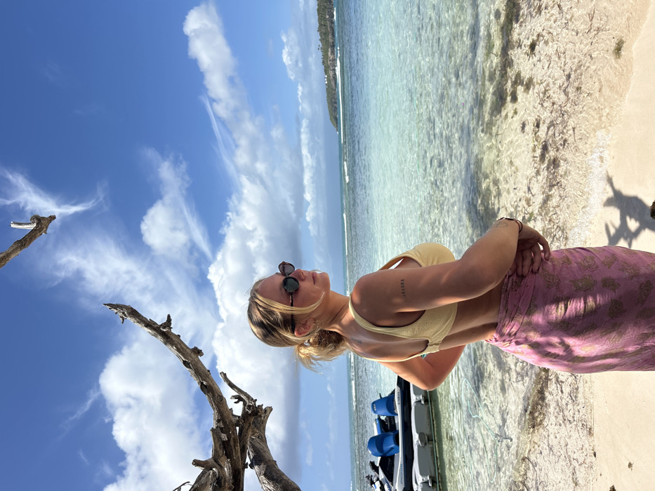 The image portrays an adult female standing on a sandy beach adjacent to shallow, clear turquoise water under a bright blue sky with scattered white clouds. This scene is located in Sainte-Anne, France. The female, appearing to be in her 20s-30s, has blonde hair pulled back into a ponytail and wears dark, oval-shaped sunglasses. She is dressed in a light yellow textured bikini top and a pink sarong with a subtle patterned design tied around her waist. A tattoo, appearing to read "SOULMA," is visible on her upper left arm. She stands with her body angled slightly right of the camera, looking towards the right side of the frame, with her right hand on her hip. To the left of the female, partially in the water, is a white and grey jet ski equipped with blue seating. A gnarled, bare wooden branch extends from the left foreground into the upper frame. The clear water reveals a sandy bottom with patches of dark marine vegetation. A tree-covered landmass is visible in the distant background, bordering the horizon line. Shadows of the female and the branch are cast on the sand, indicating bright overhead sunlight.