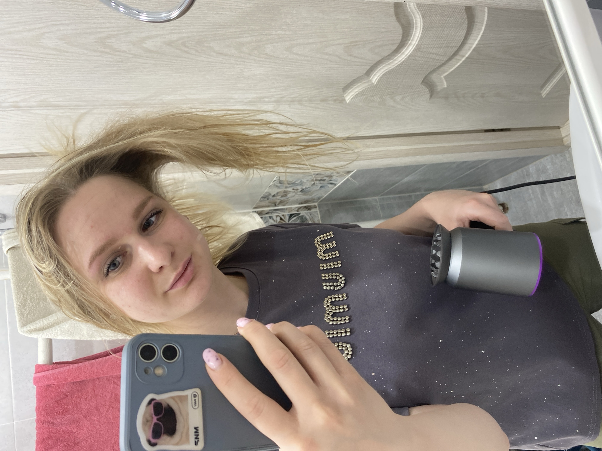 A woman is taking a selfie with her phone while holding a hairdryer. She has long blonde hair, and is wearing a grey t-shirt with a beaded design. The phone has a sticker of a pug wearing sunglasses on the back. The background appears to be a bathroom, with a towel rack and tiled walls visible. The location is Unknown, Unknown.