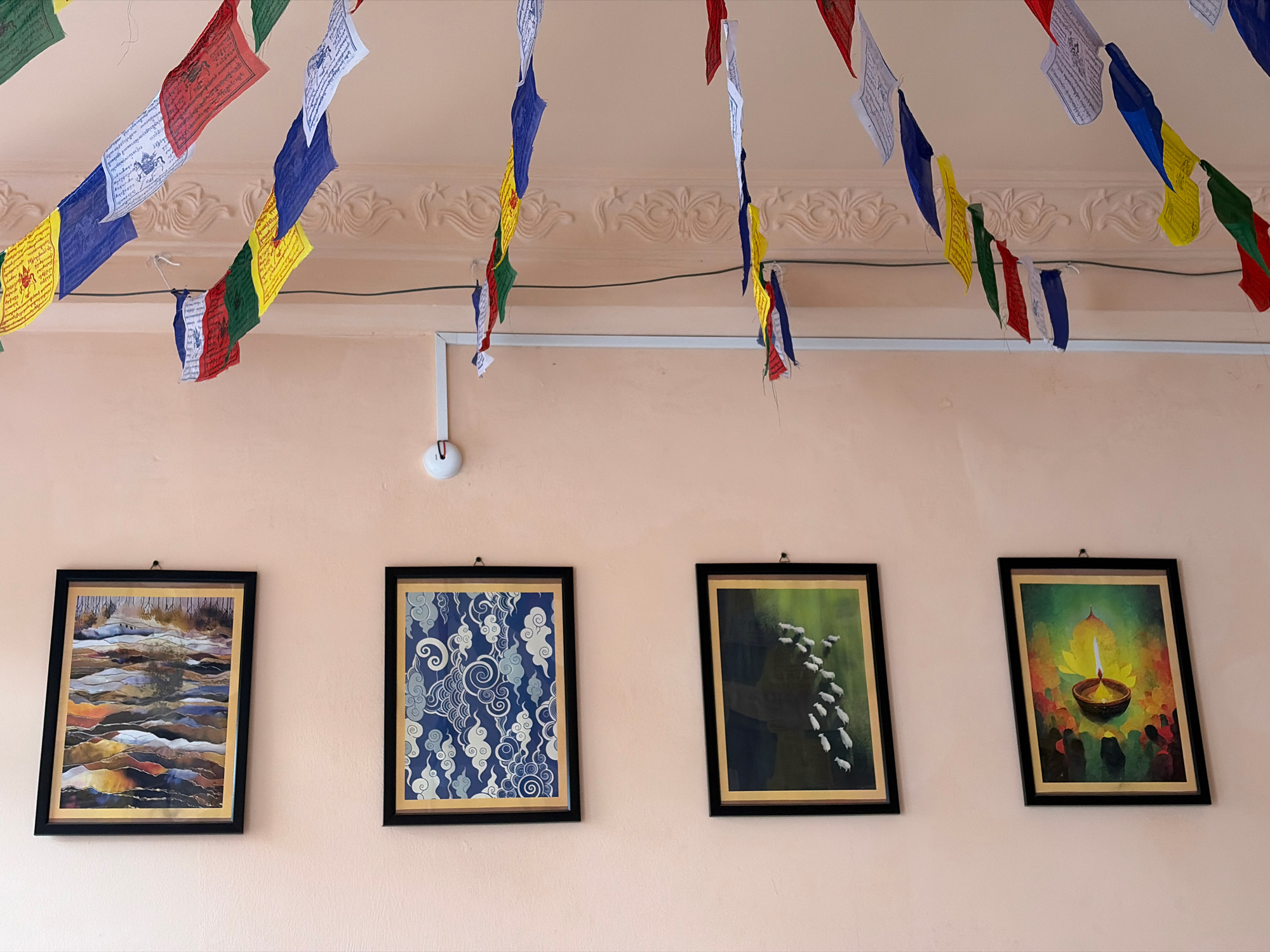 The image depicts a wall in what appears to be a room in Lalitpur, Nepal. Prayer flags are strung across the ceiling, displaying traditional Tibetan Buddhist motifs and colors. Four framed pictures hang on the light peach-colored wall. From left to right, the first picture is an abstract landscape with earthy tones. The second features a blue background with white swirling cloud-like patterns. The third shows a flock of white sheep or goats silhouetted against a dark green background. The fourth, and final picture, displays a lit diya (oil lamp) with a yellow lotus flower behind it, surrounded by dark figures. The overall impression is of a decorated interior space, possibly a home or a cultural center, in Lalitpur.