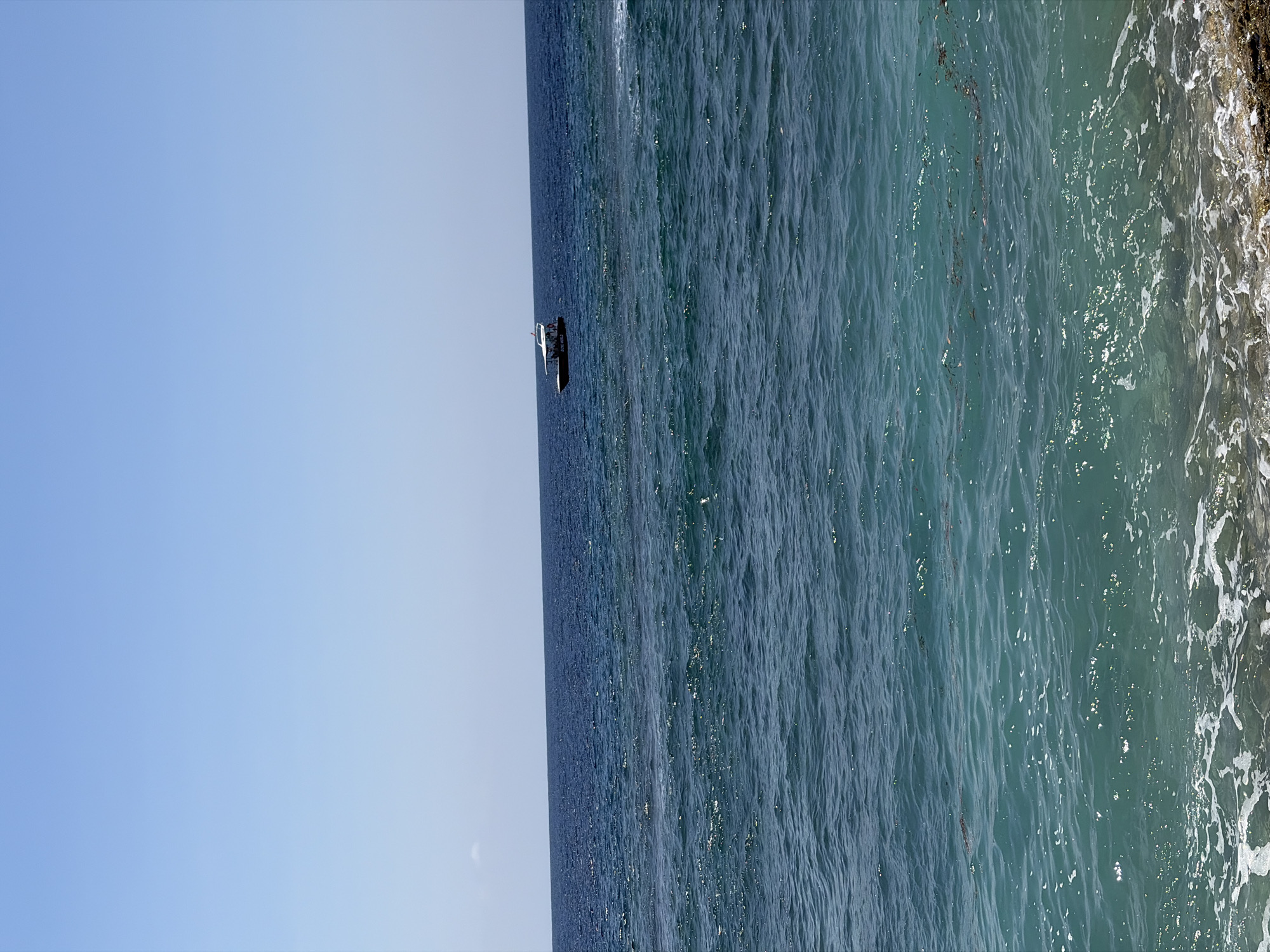 A wide-angle, eye-level shot captures a serene seascape with a clear blue sky above a vast expanse of the ocean. In the distance, a small boat with a white canopy and several passengers is visible on the dark blue water. The foreground displays the turquoise waters closer to the shore, with gentle waves lapping against a rocky coastline. The image is likely taken in Nusa Penida, Indonesia, a location known for its beautiful beaches and ocean views.