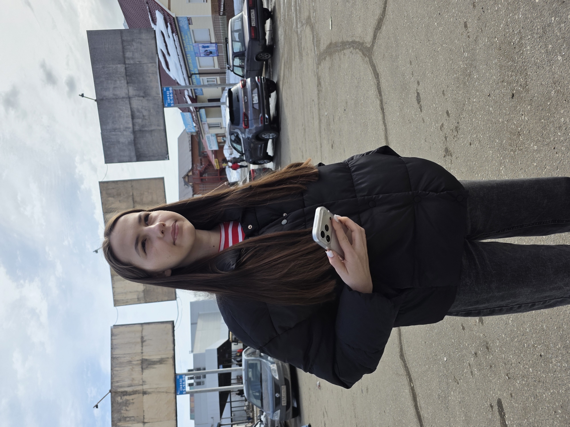 A young woman stands outdoors on a paved surface in what appears to be a city environment. She is wearing a black puffer jacket over a striped shirt and dark-colored jeans. She holds a silver smartphone in her right hand. Her long, brown hair falls over her shoulders. The background includes several cars, buildings, and billboard-like structures. Based on visual cues such as signage in Cyrillic, and previously learned knowledge, the location is determined to be within Votkinsk, Russia. The sun illuminates the scene, suggesting it is daytime.