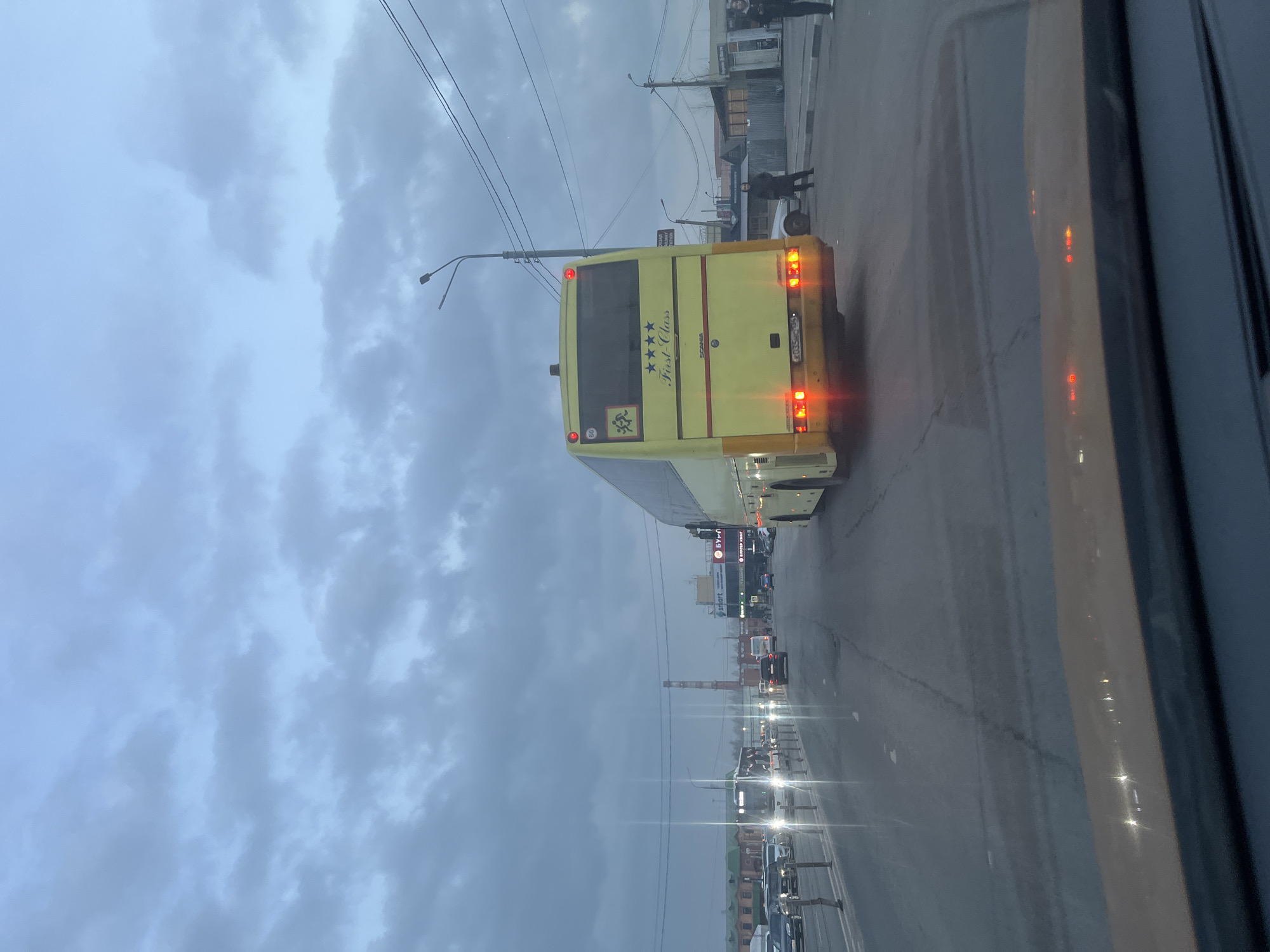 The image shows the back of a yellow "First Class" bus in Klin, Russia. The bus has its brake lights on. There are pedestrians on the sidewalk to the right of the bus. Other vehicles are visible in the distance on the road. The sky is overcast. The image appears to be taken from inside another vehicle.