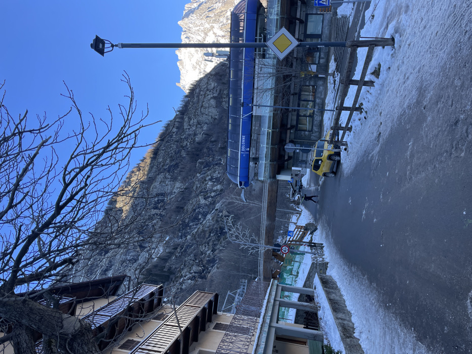 A vibrant winter scene in Courmayeur, Italy, depicts a clear, sunny day in a mountain town. The image, oriented as if viewed looking up a street towards a mountain, features a steep, rocky mountain face dominating the background, with sunlit, snow-capped peaks visible further up.
In the mid-ground, a prominent blue gondola station, labeled "DOLONNE," runs parallel to the street, suggesting a ski resort or mountain transport hub. Below it, a paved road, partially covered with snow and ice, stretches from the foreground into the distance. A yellow jeep-like vehicle is parked on the right side of the road, and a single person walks along the icy pavement in the mid-ground. A wooden fence lines the roadside.
To the left, a bare-branched tree occupies the immediate foreground, its intricate branches silhouetted against a brilliant clear blue sky. Partially visible buildings with balconies, typical of alpine architecture, are located on the left and right sides of the foreground and mid-ground.
Notable objects include a street lamp, a yellow diamond-shaped road sign indicating a priority road, a blue square sign for a pedestrian crossing, and a circular sign with a "2.1m" height limit. The clear sky and strong shadows suggest it is daytime, likely morning or early afternoon, with cold, crisp weather conditions conducive to snow and ice. The overall setting is that of an active, functional mountain resort town during the winter season.