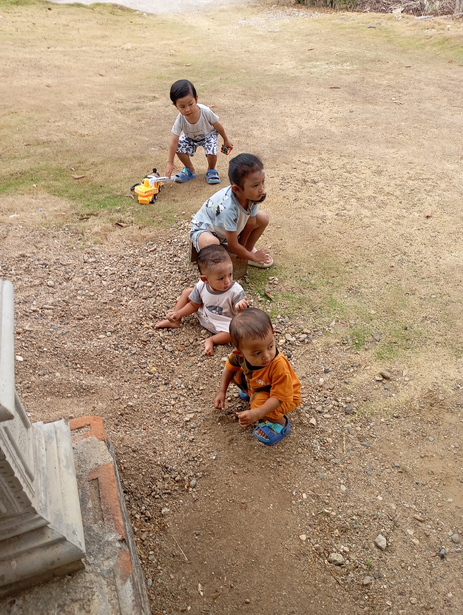 Four young children are playing on a gravel and dirt area near dry grass. The children are positioned in a line, with the youngest in the front. A boy wearing an orange shirt and blue sandals is squatting and reaching towards something on the ground. Next to him, a baby in a light-colored onesie is sitting on the ground. Further back, another child in a light blue shirt is crouched down. The furthest child, wearing a gray shirt and blue shorts, is standing and interacting with a yellow toy truck. The scene appears to be outdoors in a casual setting, possibly a residential area in Ketepung, Indonesia, given the presence of dry grass and gravel. There are also stairs visible on the left side of the frame.