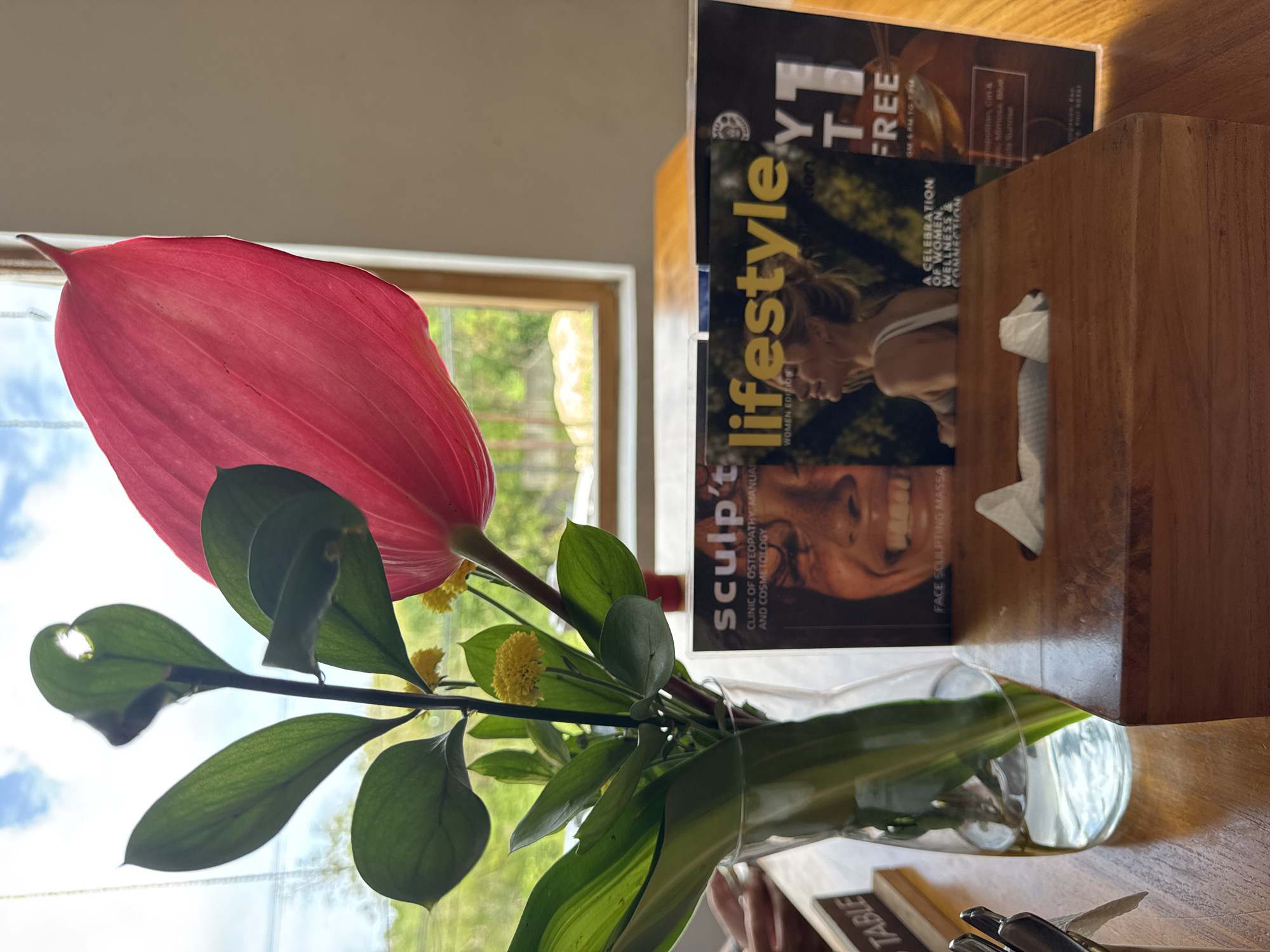 A close-up shot captures a wooden tissue box on a polished wooden table, with a glass vase holding a tropical flower arrangement to its left. The flower boasts a large, bright pink spathe and smaller yellow blooms, set against a backdrop of green foliage and a window overlooking an outdoor scene with lush greenery and a cloudy sky. Two magazines are partially visible behind the tissue box, one with the title "sculpt't" and another titled "lifestyle" featuring a woman on the cover. To the far left, a small wooden sign reads "TABLE." The overall setting suggests an indoor dining or waiting area, possibly a cafe or spa, with natural light illuminating the scene. The image is shot from a slightly elevated perspective, focusing on the foreground arrangement.