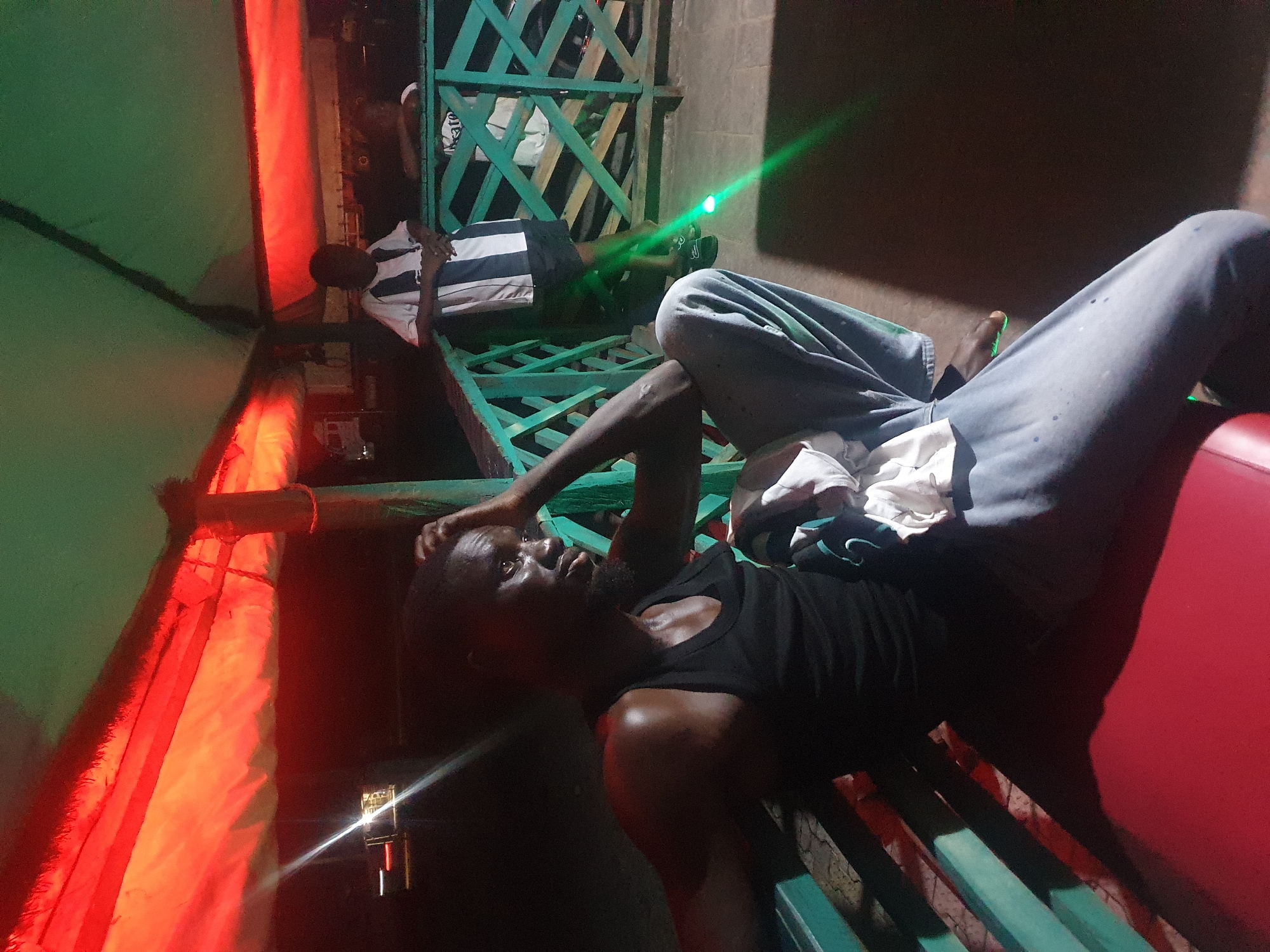 In Lokoja, Nigeria, a man wearing a black tank top and grey pants sits on a red bench under a canopy illuminated by red and green lights. He is seated on a bench with a green railing, his hand resting on his head as he looks to his right. To his right, another person wearing a striped shirt stands against the railing. The setting appears to be an outdoor relaxation area with a tiled floor.