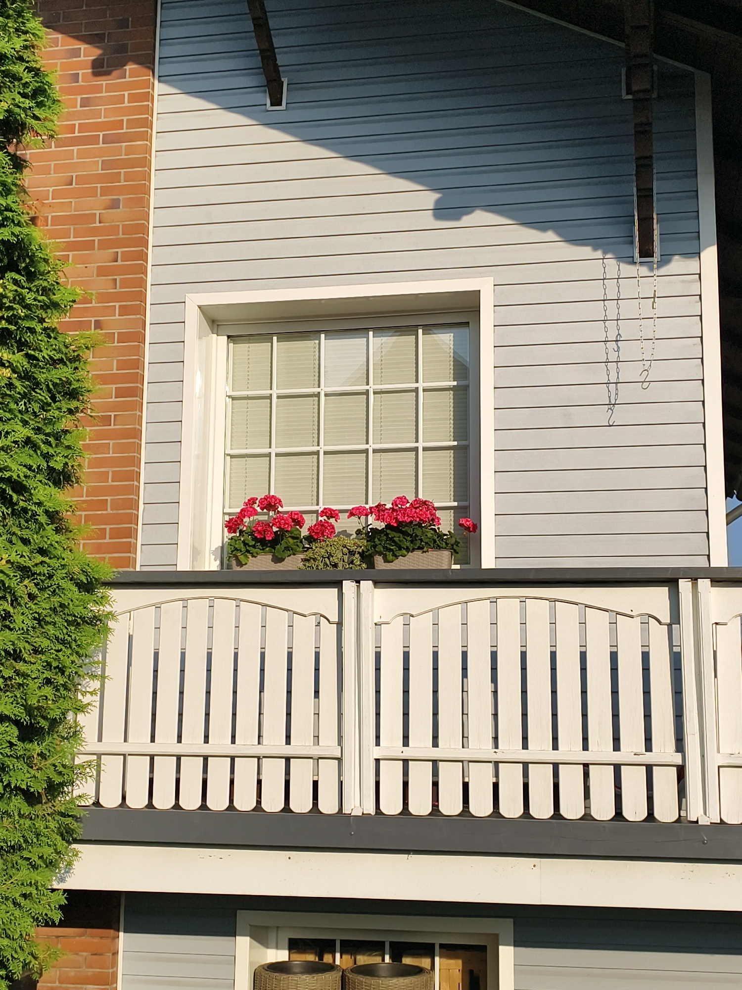 The image shows a section of a multi-story building in Tczew, Poland. The building's facade features a combination of light blue horizontal siding and red brickwork. A prominent white-framed window with multiple panes is visible, adorned with window boxes containing red geraniums. Below the window is a white picket fence that encloses a balcony or porch. A lush green evergreen tree partially obscures the left side of the frame. The scene is bathed in sunlight, casting shadows on the building's siding, suggesting it is daytime. There are no people or animals present in the image. The architecture appears residential or possibly commercial.