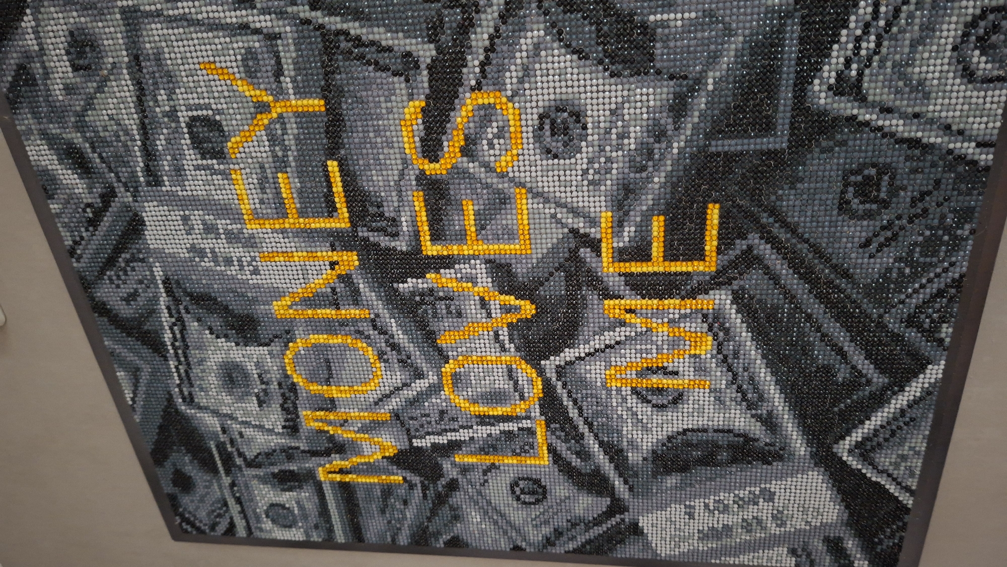 The image shows a pixelated artwork on display. The background consists of grayscale dollar bills with a mosaic effect. The phrase "MONEY LOVE ME" is overlaid in bright yellow pixelated lettering. The artwork is framed with a dark gray border. The photograph's angle suggests it was taken indoors, with a plain wall visible in the background. There is no evidence to suggest that the photo was taken in Chaikovsky, Russia.