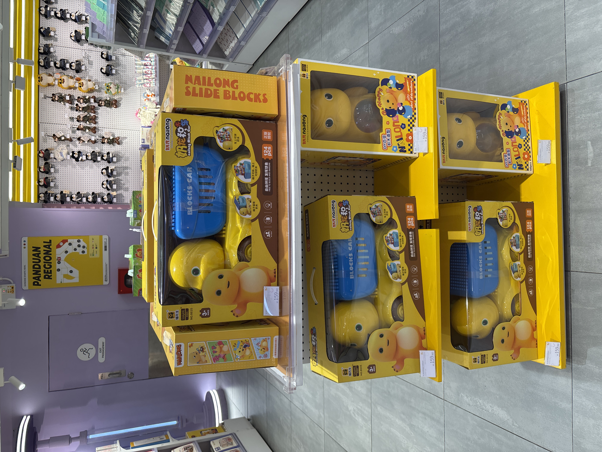 The image shows a retail display of toy sets in what appears to be a store in West Jakarta, Indonesia. The primary focus is on a shelf of "nailong" branded toy sets. The toy sets are mostly yellow and blue, featuring cartoon dinosaur-like characters and building blocks, with text in Chinese characters on the packaging. The prices of the toy sets are marked as 159,900 Indonesian Rupiah.
In the background, there's a shelf with plush toys and a lilac-colored wall with a sign in Indonesian that says "Panduan Regional", an exit sign with a pictogram, and a door. The store has tiled flooring and artificial lighting. No people are clearly visible.