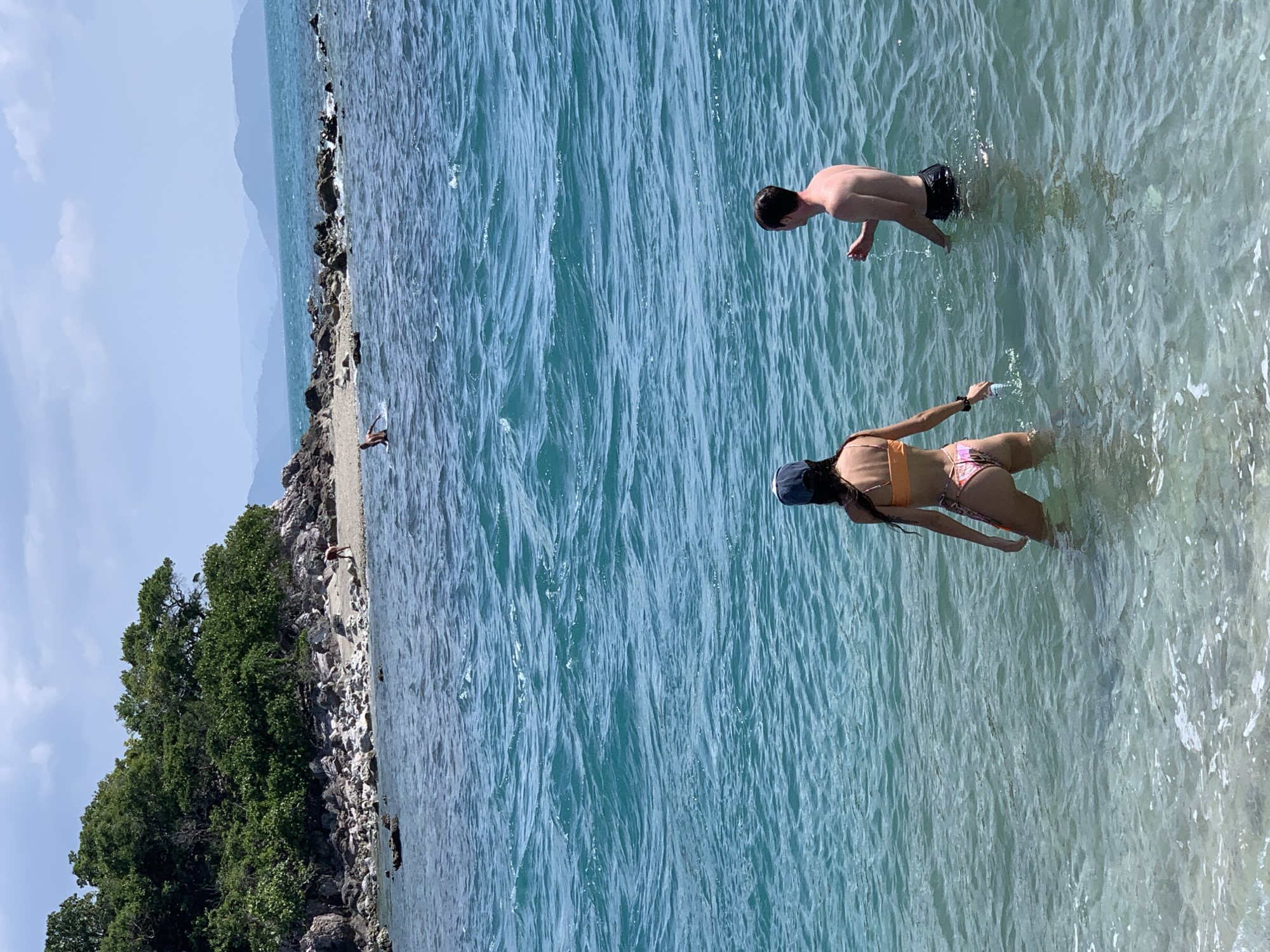 In a turquoise ocean, two individuals are wading in the water. A woman, facing away from the camera, wears an orange bikini and a dark baseball cap. A man, also facing away, has dark hair and wears black swim trunks. In the background, a rocky island with lush green vegetation rises from the sea. Further in the distance, hazy mountains form the horizon under a bright, partly cloudy sky.