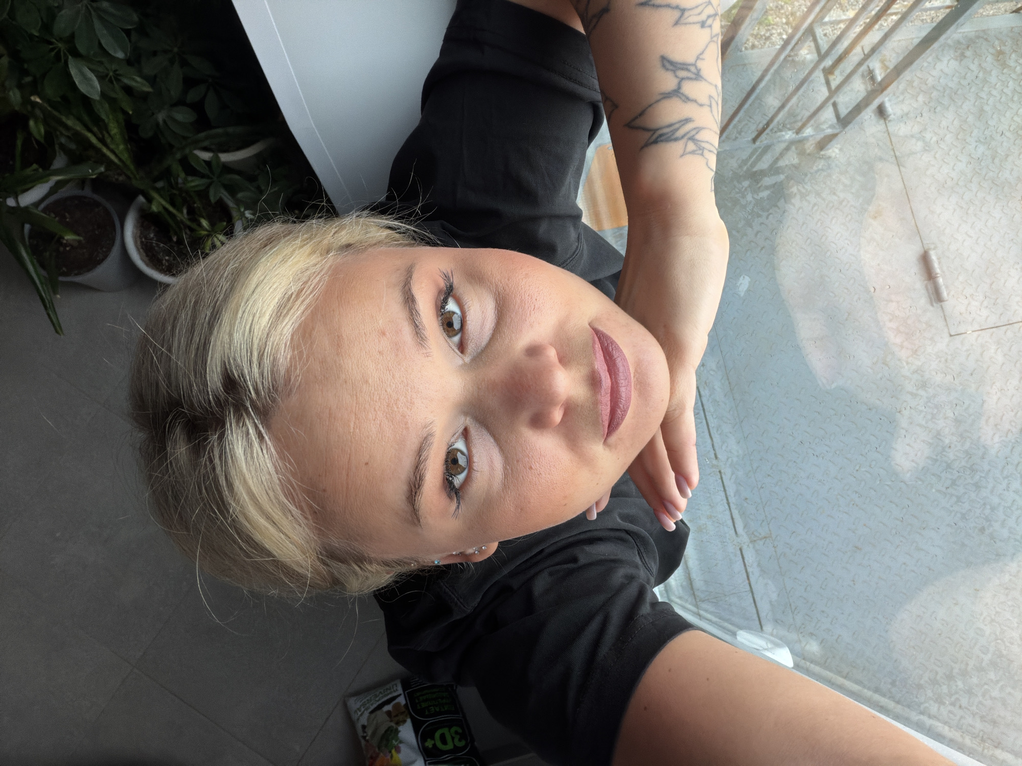 A young woman is captured in a high-angle selfie, looking directly up at the camera with a neutral expression. She has blonde hair, light skin, and striking brown eyes. Her left arm, visible in the foreground, is adorned with a tattoo featuring linear designs, and she wears a black short-sleeved top. Multiple ear piercings are visible on her left ear, and her fingernails are manicured with a light polish.
The setting appears to be an indoor or covered balcony space in Sarapul, Russia. To the left, several potted green plants provide a natural element against a dark background. Below her, the floor is a dark grey tiled surface. On the bottom left, a product bag with Cyrillic text is visible, clearly showing "3D+" along with words like "УНИВЕРСАЛ" (UNIVERSAL) and "ГРУНТ" (SOIL). To the right of the woman, a window or balcony opening reveals a light-colored metal grate or diamond plate surface outside, with a white frame or railing. The scene is brightly illuminated, indicating daytime.