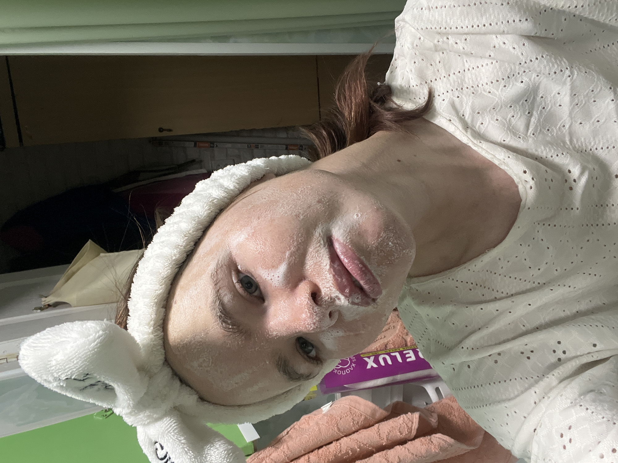 A woman is posing for a selfie in what appears to be an indoor setting, possibly a bathroom, based on the presence of cabinets and a countertop. She is wearing a white, eyelet-patterned top and a white terry cloth headband with bunny ears. Her face is covered in a white substance, likely a face mask or cleanser. There's a pink towel and a purple box with "RELUX" written on it nearby. The location is Chaikovsky, Russia.