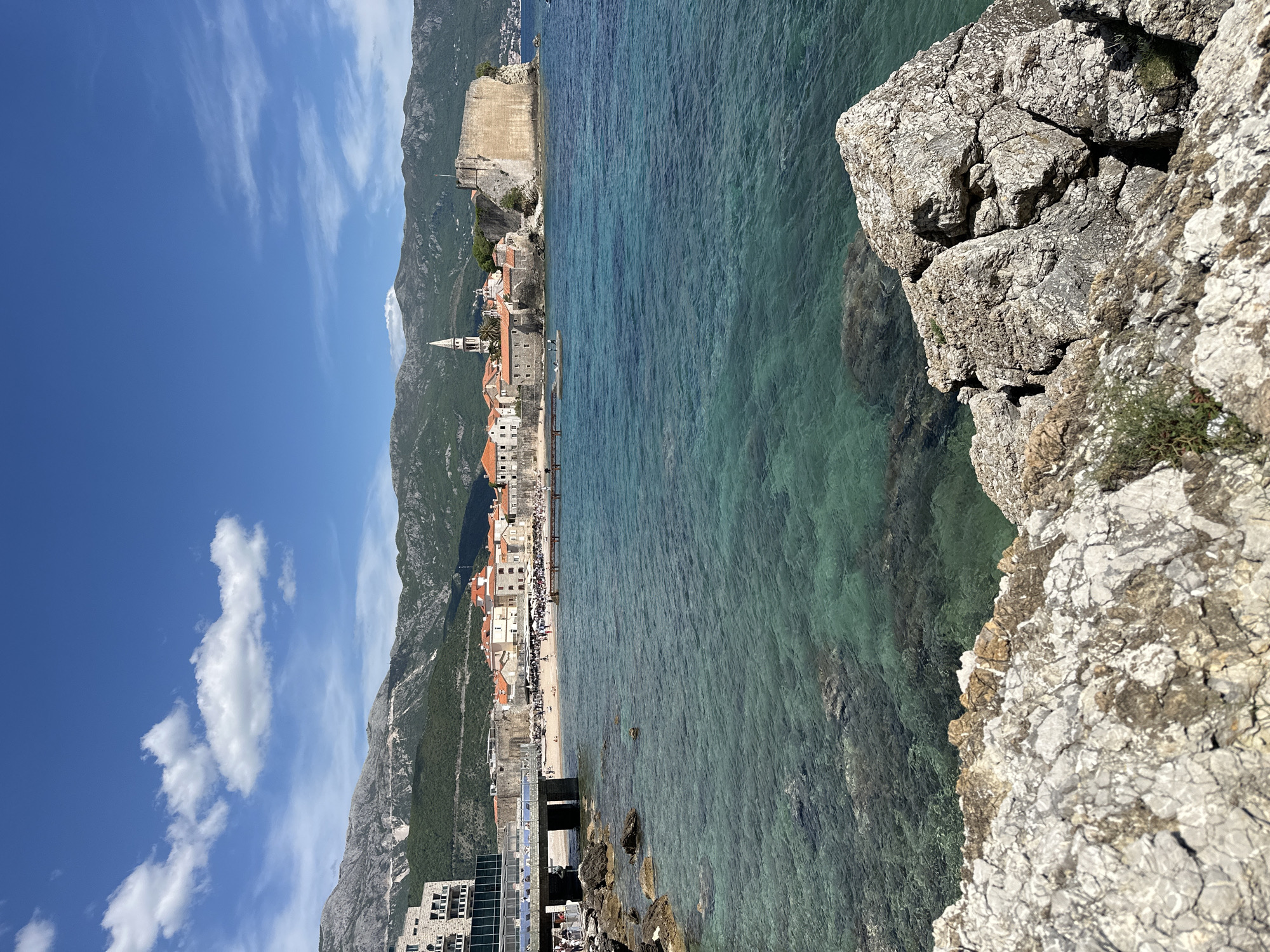 The image depicts a coastal scene in Budva, Montenegro, taken from an elevated perspective. The Adriatic Sea is visible in varying shades of turquoise, with rocky outcrops along the shore and beneath the water's surface. The medieval old town of Budva is seen along the coastline, with its characteristic red-tiled roofs and historic buildings, including a prominent church tower.
Mountains form a backdrop to the town, with lush green vegetation covering their slopes. Above, a blue sky is scattered with white clouds. In the foreground, a rocky cliff face dominates the view, composed of gray stone with visible cracks and textures. The positioning of the viewpoint suggests the photo was taken from a high point near or along the edge of the coastal cliffs surrounding the town.