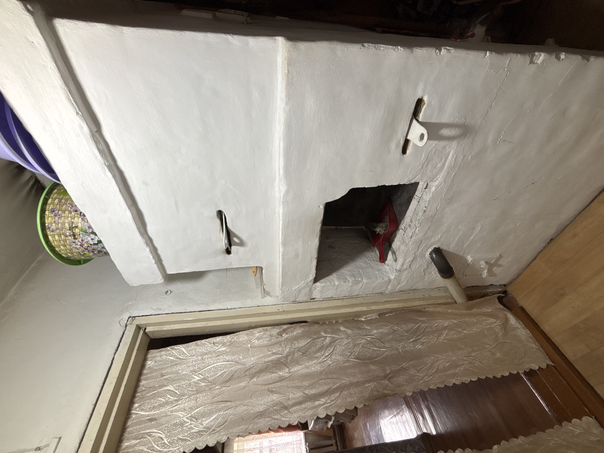 The image depicts the interior of a room, likely a kitchen or living area, in a dwelling in Ilpanur, Russia. The primary focus is a large, white, traditional Russian stove (pech) dominating the right side of the frame. The stove is constructed of plaster and appears to be functional, with visible openings and metal hardware, including what looks like a damper or vent. There's a small red shovel with a brush inside one of the stove's openings, suggesting recent use.
To the left of the stove, a doorway is visible, partially obscured by a light-colored, textured curtain with a floral pattern. Through the doorway, a glimpse of another room is offered, revealing a wooden floor and what appears to be a window with a sheer curtain. The walls in the immediate vicinity of the stove and doorway are also plastered and painted white.
On top of the stove, a woven basket with a colorful floral design is placed, and partially visible behind it are stacked purple items, possibly plates or lids. A section of the wall above the stove has a visible crack or repair. A white pipe emerges from the wall near the base of the stove and extends horizontally. The floor is covered with light-colored wooden planks. The overall impression is of a rustic, traditional living space.
