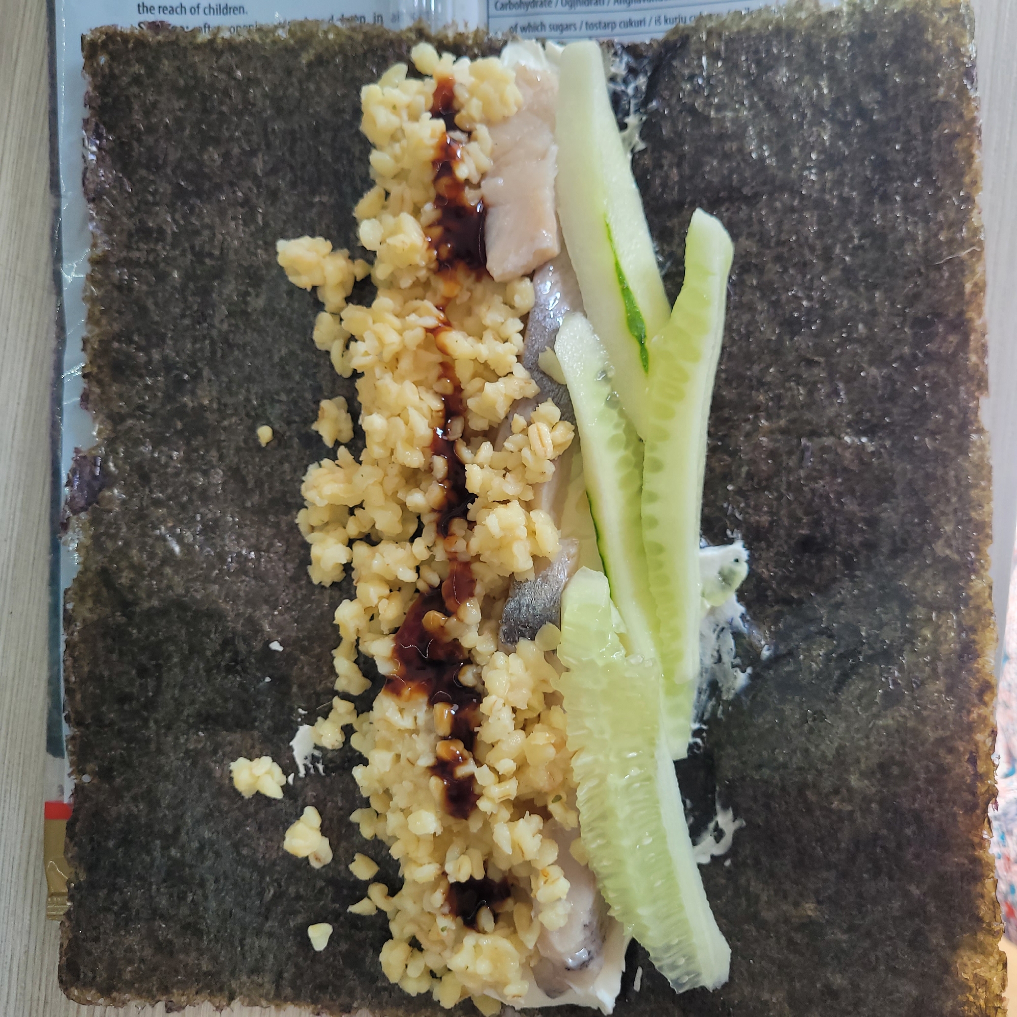 The image shows a close-up overhead view of a piece of nori seaweed on a wooden surface, presumably in a kitchen. On the nori, rice, pieces of fish, sliced cucumber, and a dark sauce are arranged in a line, suggesting the preparation of a sushi roll. The background is out of focus but appears to be a wooden table or countertop. There is no indication of Jūrmala, Latvia.