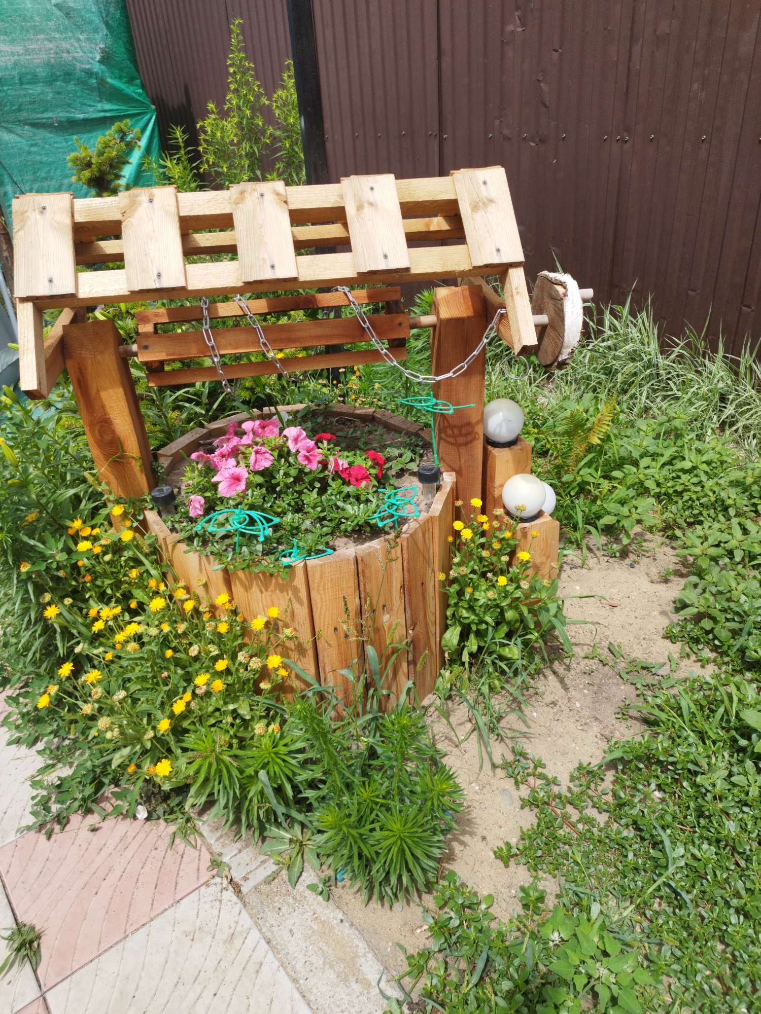 This image shows a decorative wooden well with a roof, located in Oskemen, Kazakhstan. The well is filled with pink and red flowers. Surrounding the well is lush green vegetation, including small yellow flowers and various types of grass. The well has a wooden crank and a chain running across the top. Two white spherical lights are placed on wooden posts to the right of the well. In the background, there is a dark brown corrugated metal fence and a green tarp. The ground is a mixture of sand and dirt, with some paving stones visible on the left side of the frame. The scene appears to be an outdoor garden setting on a sunny day.