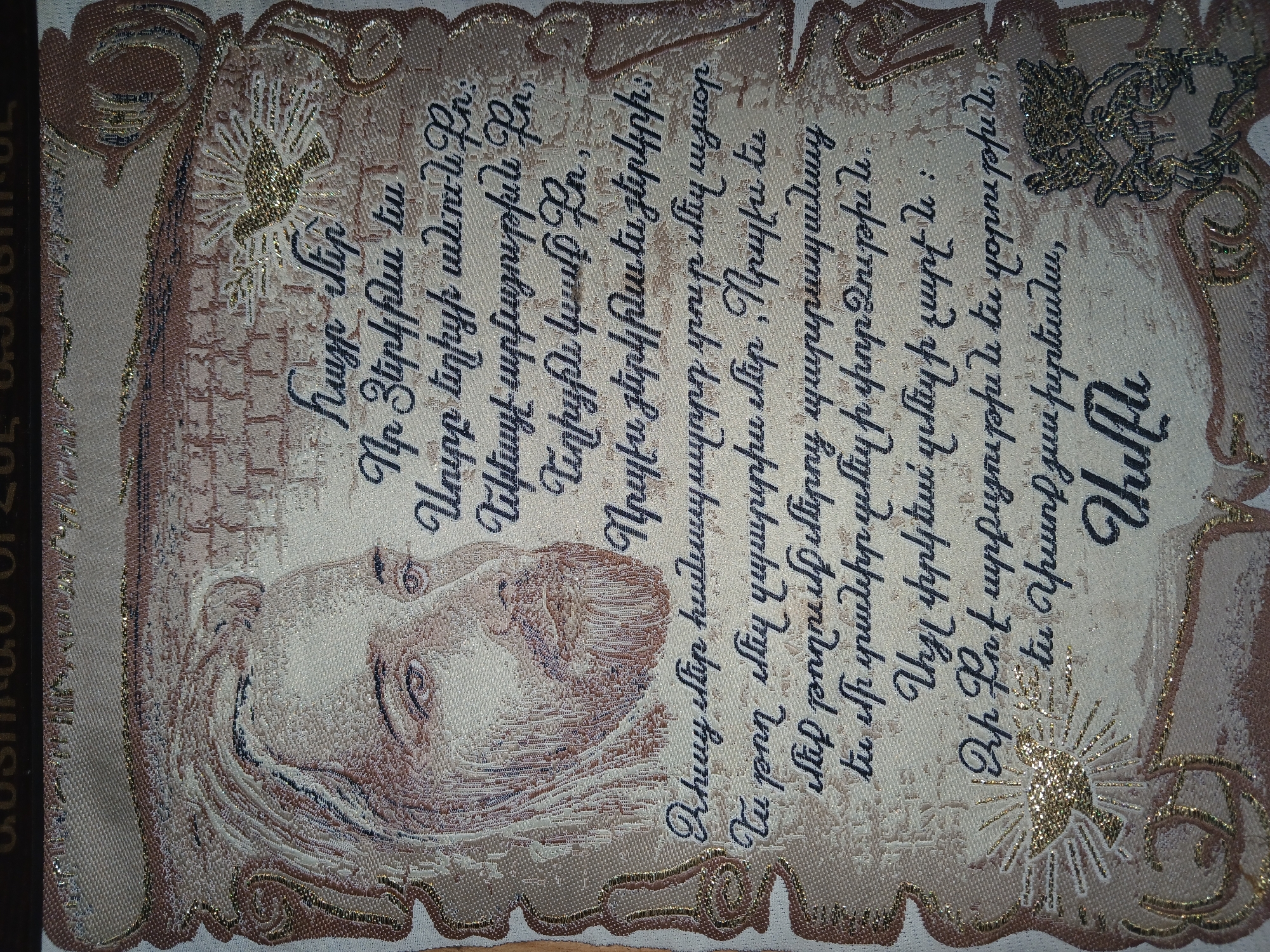 The image displays a close-up view of a tapestry or textile artwork featuring a portrait of a man with a beard and long hair, reminiscent of religious iconography, along with Armenian script and decorative elements. The man's face is rendered in shades of white, beige, and brown, with detailed brushwork or weaving techniques creating texture and depth. His eyes are looking forward. To the right of the portrait, Armenian text is prominently displayed in a dark, script-like font. Decorative motifs, including what appears to be a dove with radiating lines and ornate scrollwork, are also present, some of which are highlighted with gold thread. The overall aesthetic suggests a spiritual or religious theme. The backdrop of the artwork is a textured fabric, likely part of the tapestry itself, with subtle patterns in beige and brown tones. There is no discernible urban environment or specific location in Ijevan, Armenia visible within the frame of the image. The image focuses solely on the textile artwork.