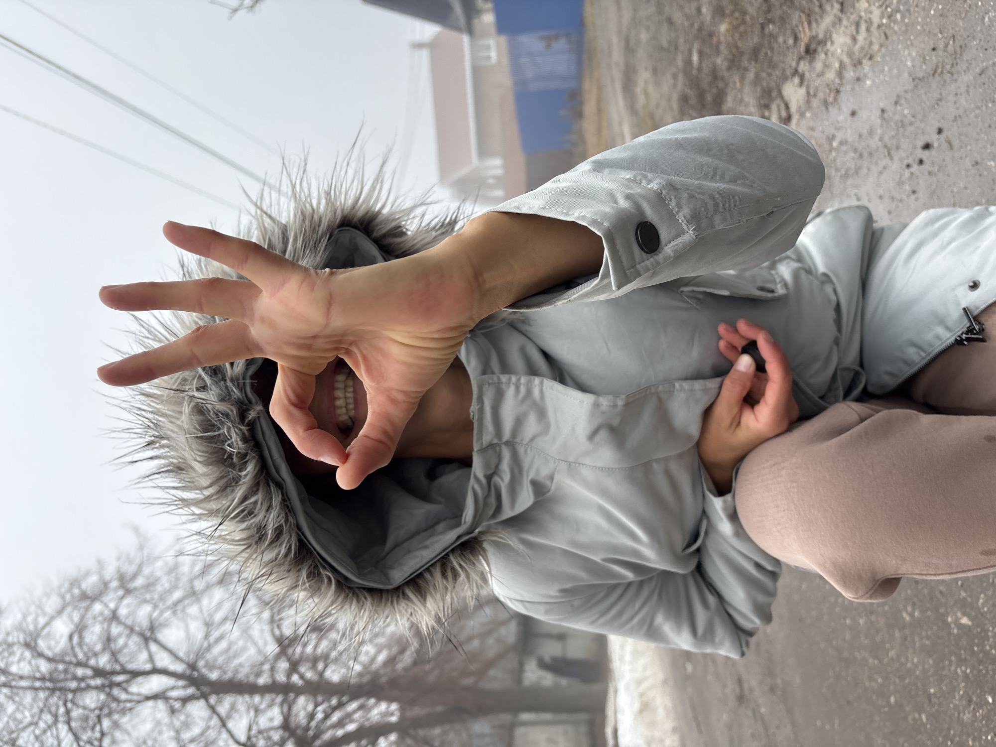 The image shows a person wearing a light grey, hooded jacket with a fur trim on the hood, standing outside on a cloudy day in Ulyanovsk, Russia. The person is holding up their left hand, making a circle with their thumb and index finger. The person's mouth is visible through the circle. They are wearing light pink pants. The ground is a concrete or asphalt surface with some patches of white, possibly snow or ice. In the background, there are buildings, trees without leaves, and a heavy fog.