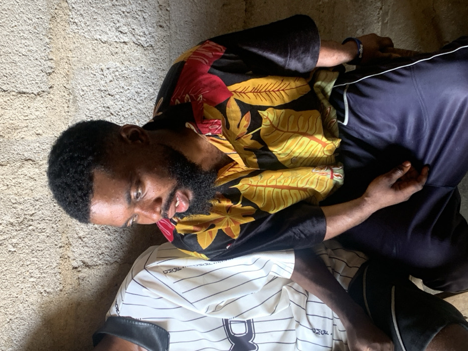 The image depicts two males seated side-by-side against a rough, light-colored concrete or plastered wall, likely within an indoor or sheltered outdoor setting in Okene, Nigeria. The prominent subject, positioned centrally, is a male with dark skin, short black textured hair with a fade, and a full black beard and mustache. He wears a short-sleeved black collared shirt featuring a pattern of large yellow and red leaves, paired with dark blue athletic shorts or trousers that have a white stripe down the side. A blue wristband with white lettering is visible on his left wrist. His mouth is slightly open, and his gaze is directed towards the right side of the frame. To his left, a second male is partially visible, wearing a white collared shirt with thin vertical black stripes and a black collar, displaying text that appears to read "OMOE" on the chest. Both individuals are seated with their hands resting on their laps. The scene is illuminated by ambient light.