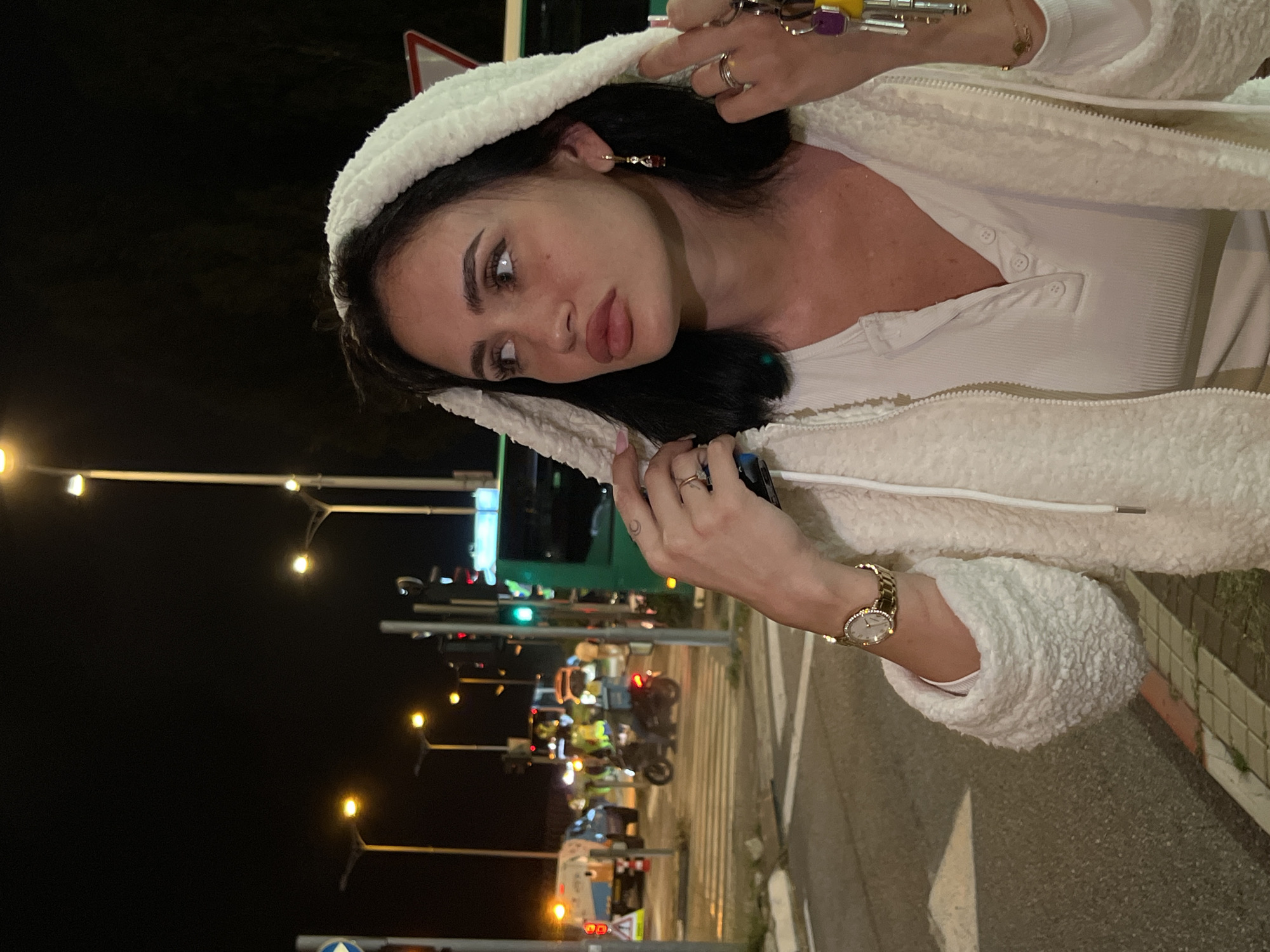 A woman with dark hair and light skin is standing near a street in Mate Asher Regional Council, Israel, at night. She is wearing a white, fluffy hooded jacket over a light-colored top. The woman is holding her hands up near her face, displaying rings on her fingers. A gold watch is visible on her left wrist. In the background, there are streetlights, traffic lights, vehicles, and pedestrians on a crosswalk.