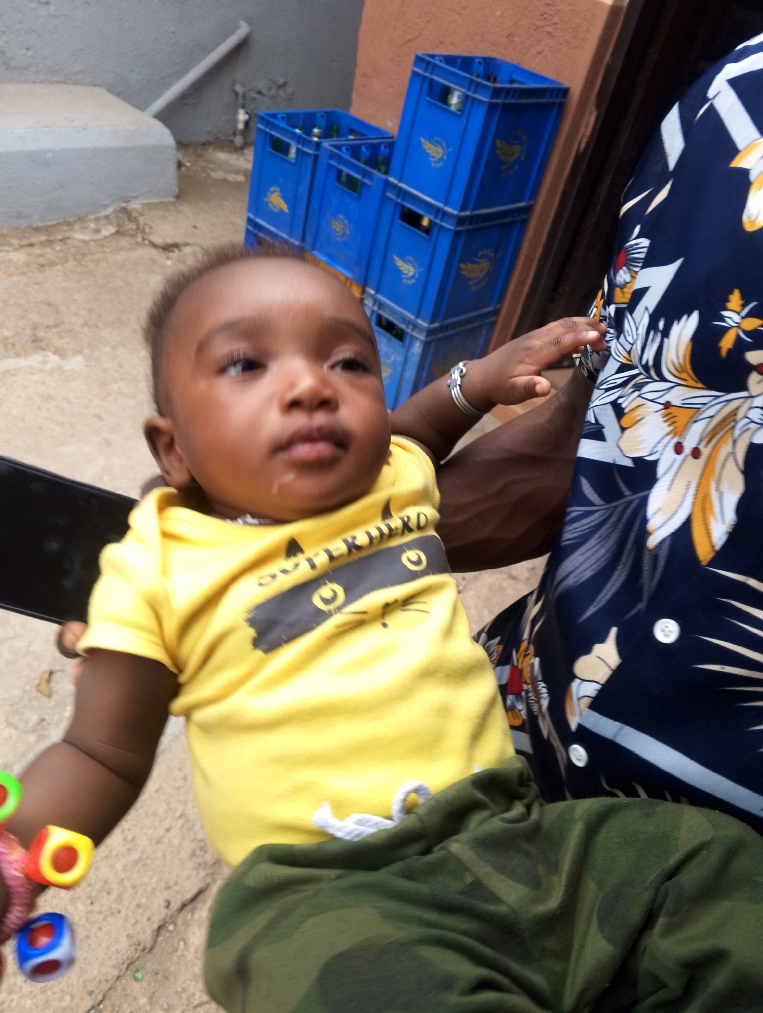 The image presents a daytime outdoor scene, likely in a paved area or courtyard in Jos, Nigeria. The primary subject is a dark-skinned baby, held by an adult whose body is partially visible on the right side of the frame.
The baby wears a bright yellow short-sleeved t-shirt featuring a black graphic with the word "SUPERHERO" partially visible, alongside a circular design. Their pants are camouflage-patterned green. The baby has short, dark hair and a neutral expression, with a small amount of drool on the chin. A silver-colored bracelet is worn on the baby's left wrist. In the baby's left hand, a colorful toy consisting of red, yellow, and blue cube-shaped beads on a string is held. A black smartphone is also visible near the baby's left side, possibly held by the baby or the adult.
The adult, whose face is not shown, is wearing a navy blue short-sleeved shirt adorned with a vibrant floral pattern of white, yellow, and orange flowers with green leaves. Their muscular, dark-skinned right arm is visible, holding the baby.
In the background, multiple blue plastic crates are stacked against a light-colored wall. These crates, which appear to contain bottles, each display a distinctive yellow bird logo. The ground is concrete, and a grey concrete step or ledge, along with a grey pipe, is visible further in the background. The natural light indicates it is daytime.