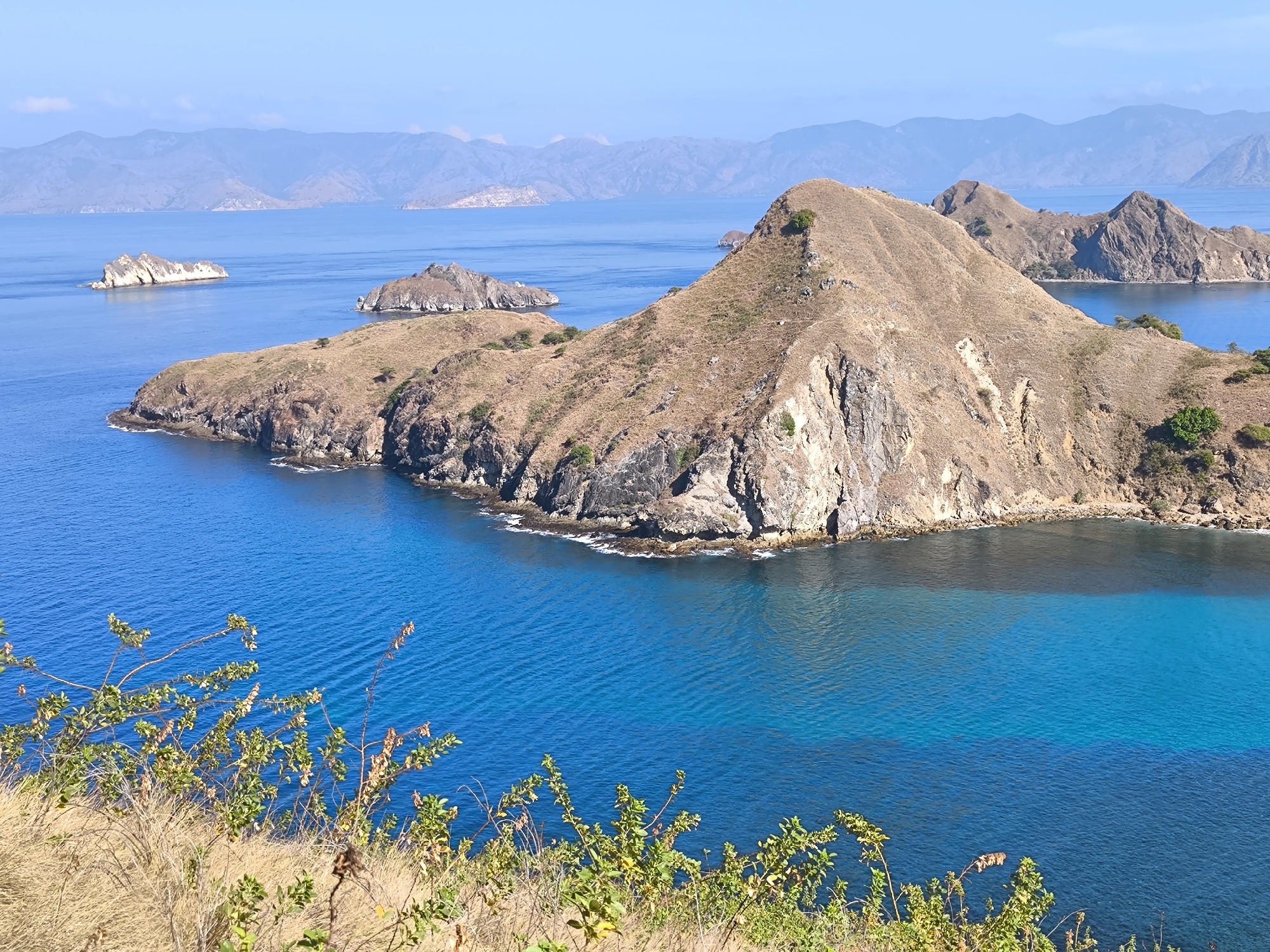 The image depicts a panoramic view of a coastline with several islands and mountains in the background. The foreground is dominated by dry, grassy hills with sparse green vegetation. The sea is a vivid blue, with a clear transition to a lighter turquoise in some areas, suggesting shallow depths or sandbanks. Several rocky islands, some with sparse vegetation, dot the water. In the distance, a chain of mountains stretches across the horizon under a clear blue sky. The overall impression is of a remote, natural landscape. The location is identified as Unknown, Indonesia.