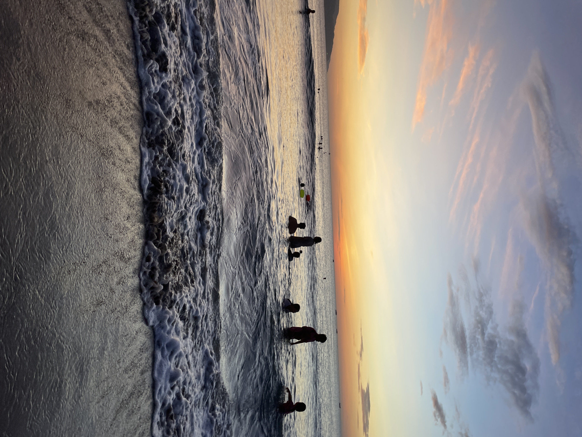 At sunset in Da Nang, Vietnam, several individuals are immersed in the ocean. The sky displays a gradient of colors, transitioning from soft pinks and oranges near the horizon to blues and purples overhead, dotted with wispy clouds. The sea is calm with gentle waves, and foam crests along the shoreline in the foreground. Silhouettes of people, including adults and children, are visible in the water, some standing, others appearing to play or swim. Small boats are scattered on the distant horizon. The overall scene evokes a serene evening at the beach.